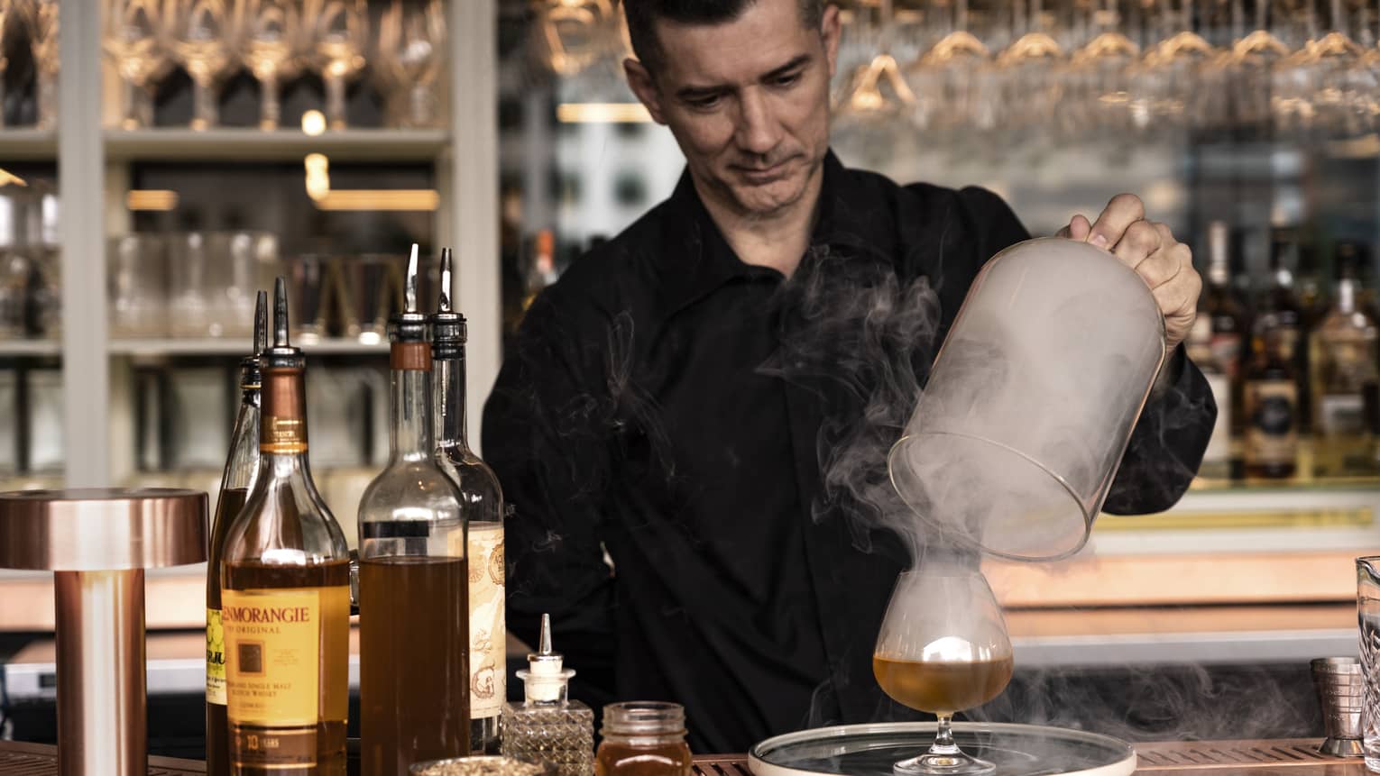 Male bartender wearing a black button-down shirt lifts up a glass bell jar revealing a cocktail glass filled with liquid and smoke