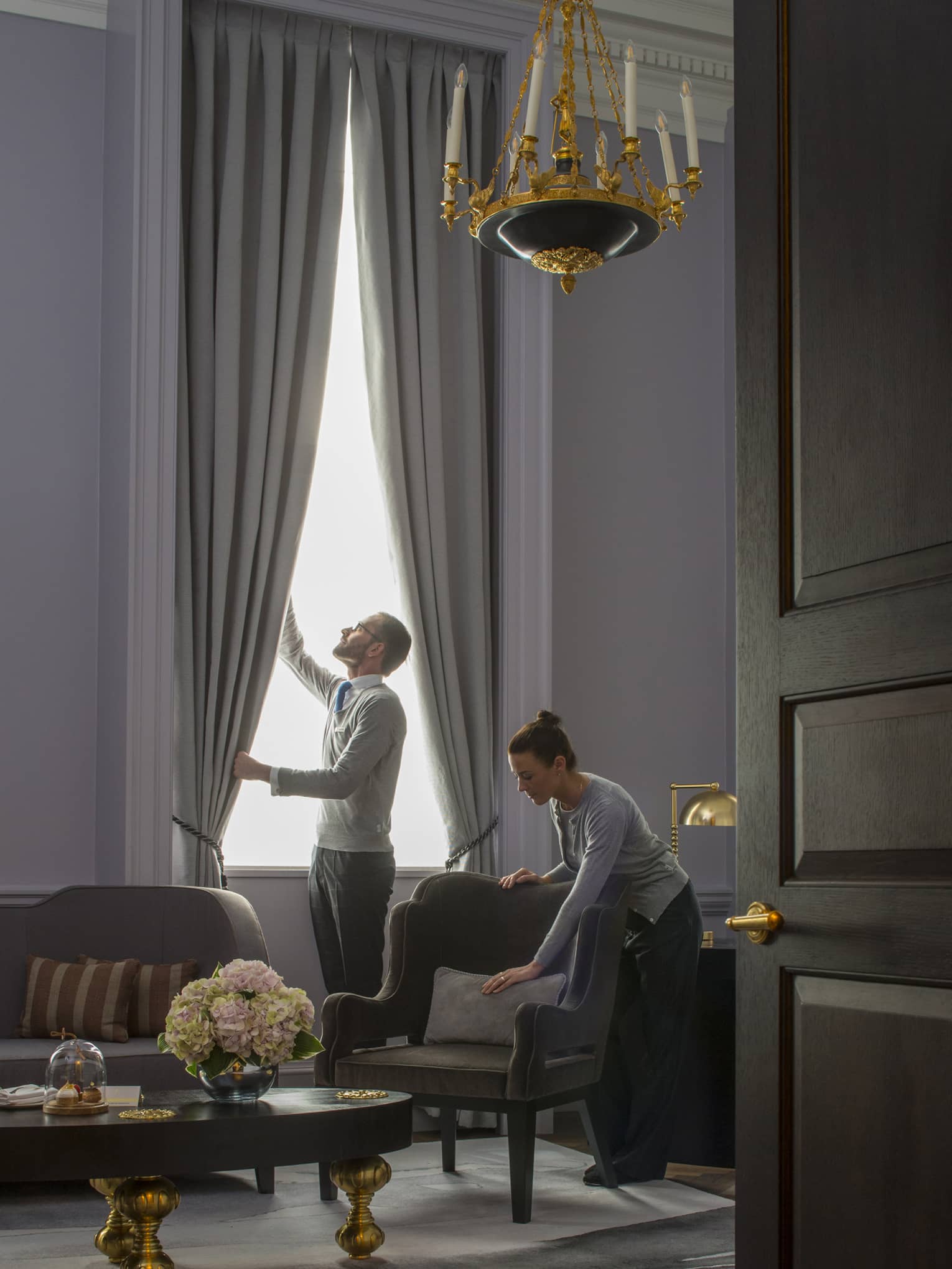 Grand Heritage Suite, man opens drapes as woman smooths pillow on armchair under chandelier 