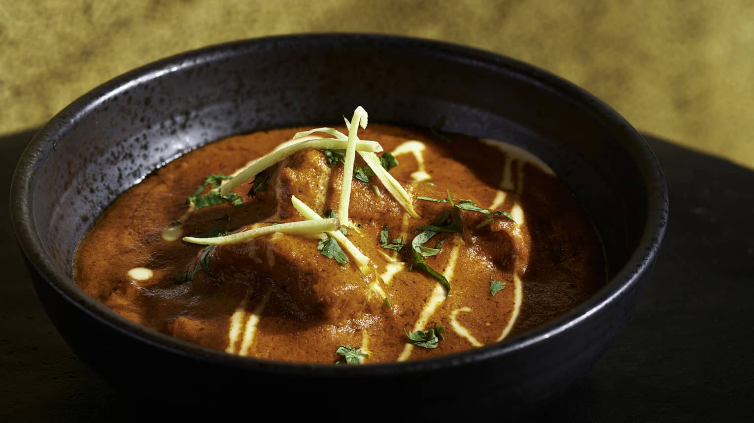 Bright red Tandoori dish with chicken, fresh herb garnish in black earthenware bowl
