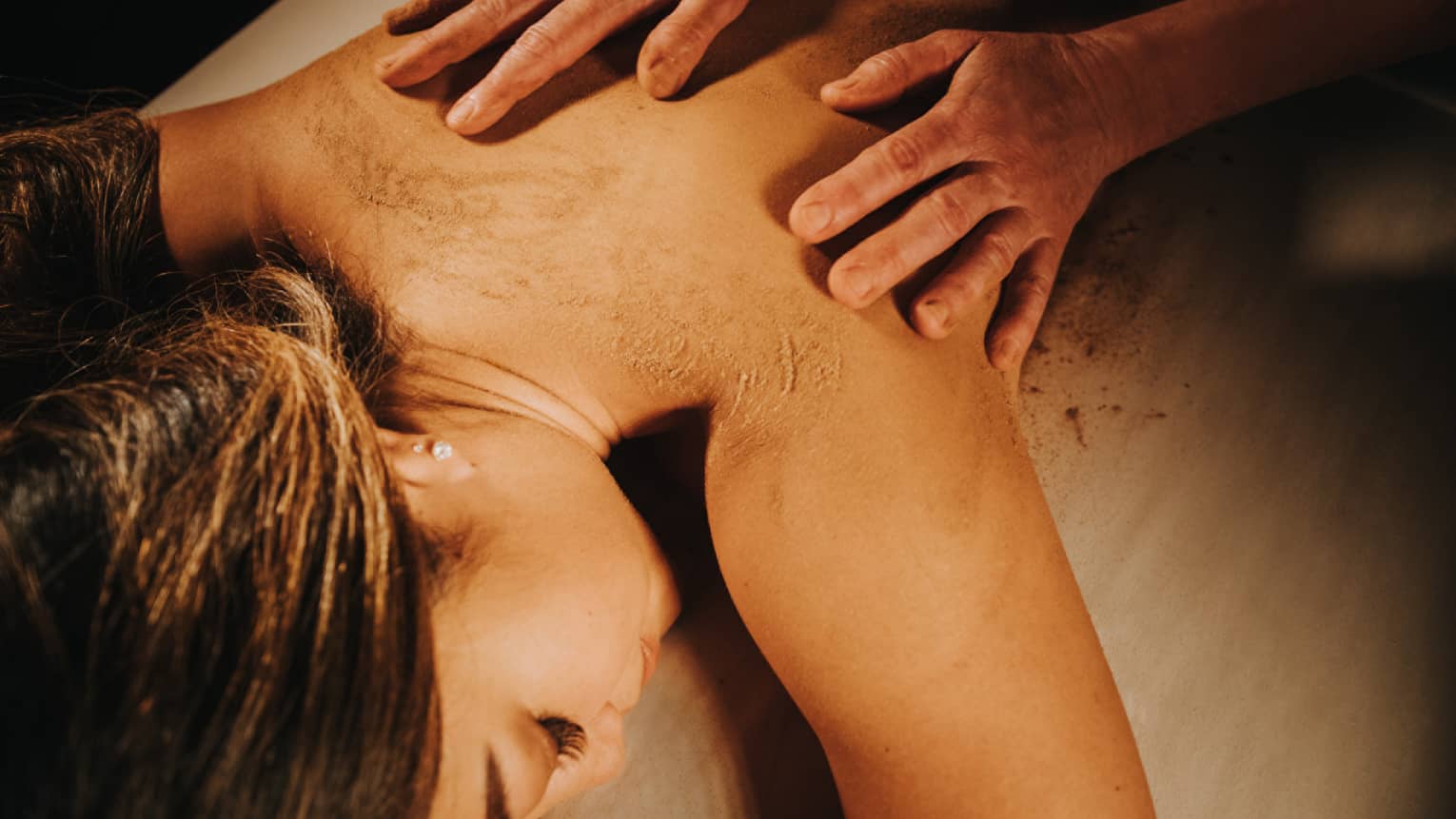 Two hands on woman's bare back as she lays on massage table in dimly lit room