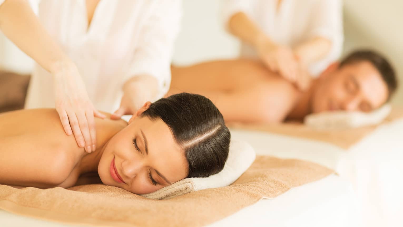 Woman and man lie side-by-side on massage tables as spa staff massage their bare shoulders
