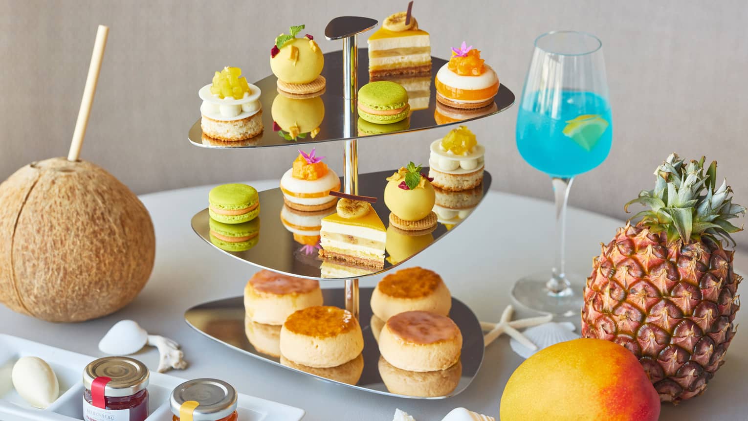 Tropical Afternoon Tea display with sweets and savouries on metal stand, whole mango, pineapple and coconut garnish