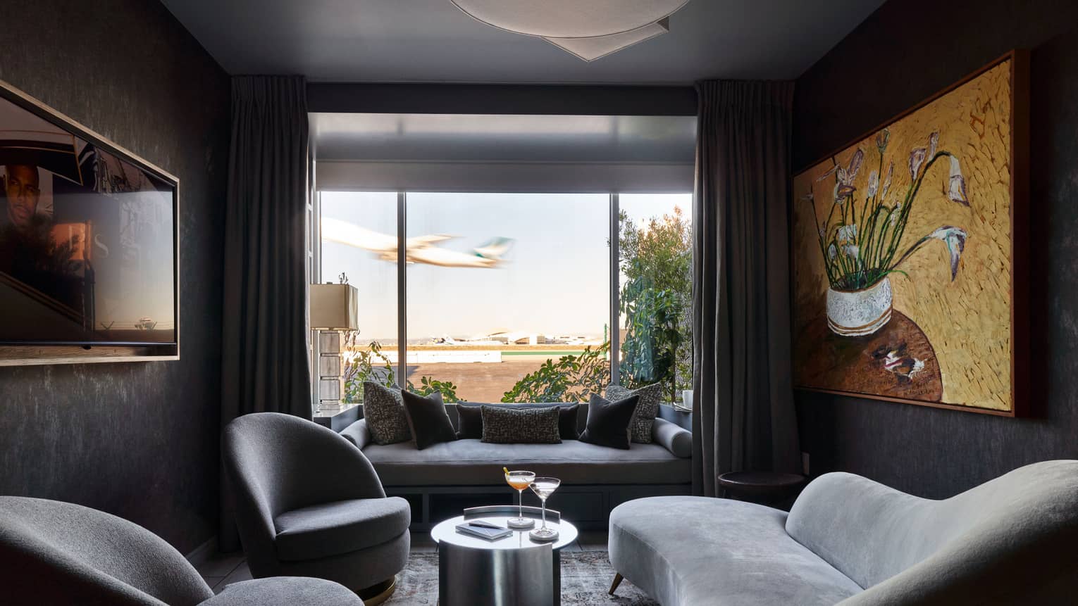 A cozy, contemporary interior space decorated in shades of grey, with a large window, plush furniture and art on the walls