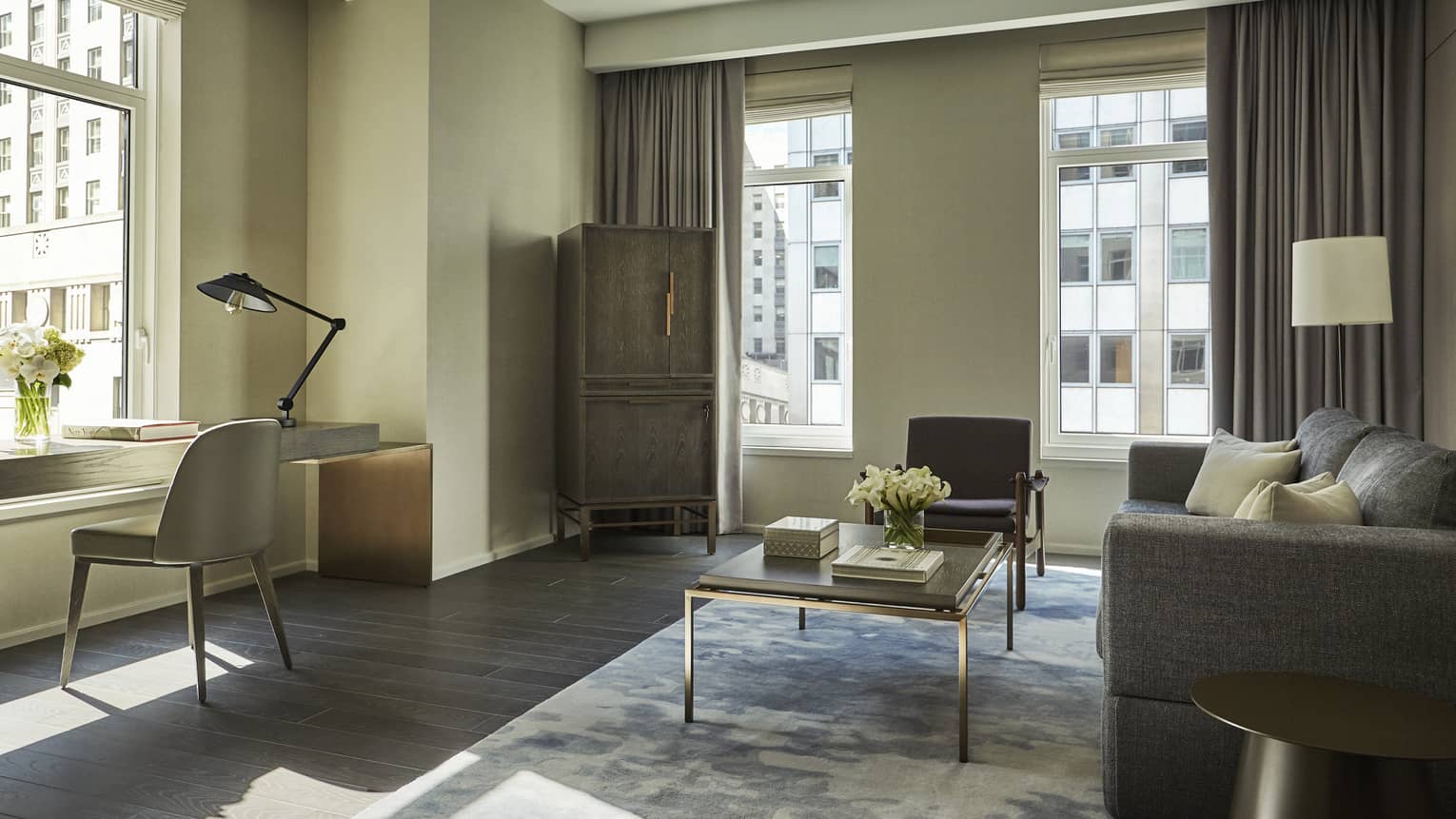 Luxury suite living room with a gray sofa, coffee table, desk with reading lamp by the window, featuring neutral tones and overlooking urban buildings
