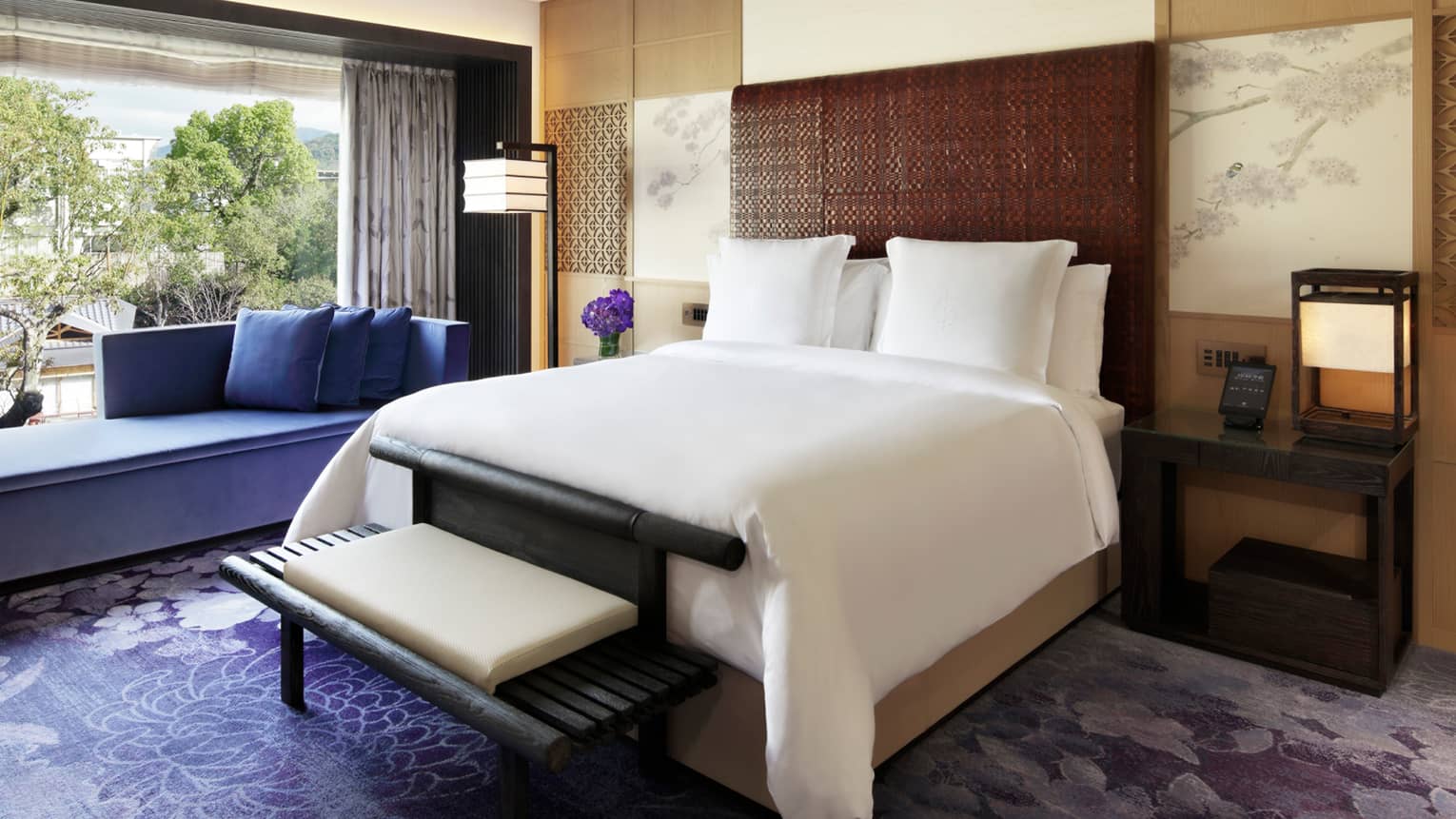 Elegant bedroom with a king bed, a purple chaise lounge, rug and flowers and floor-to-ceiling windows overlooking lush greenery