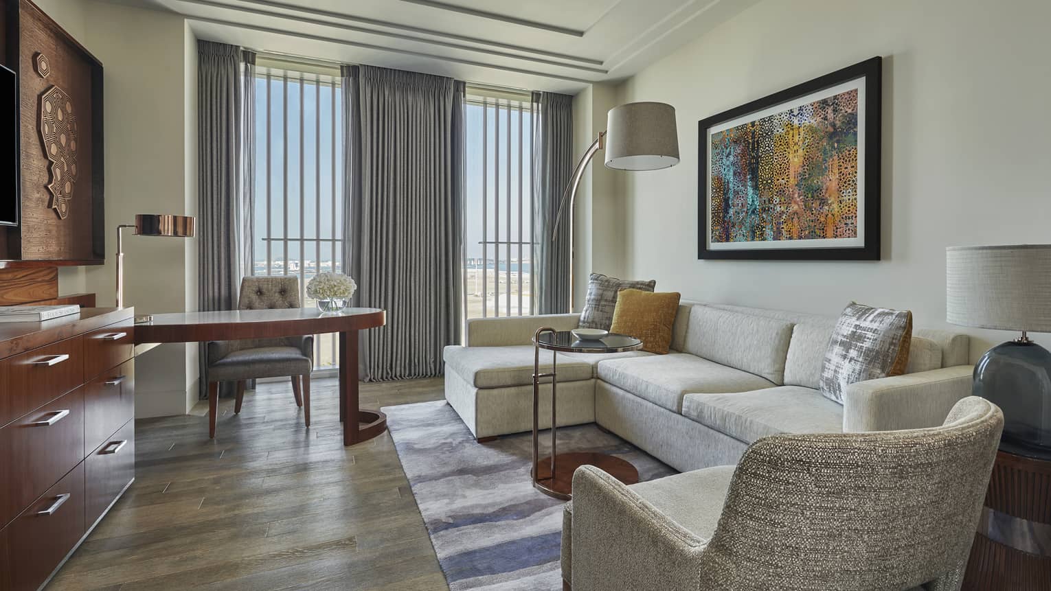 Modern hotel living area with a cream sectional sofa, colorful artwork, a wooden desk and large windows with gray curtains overlooking a scenic view
