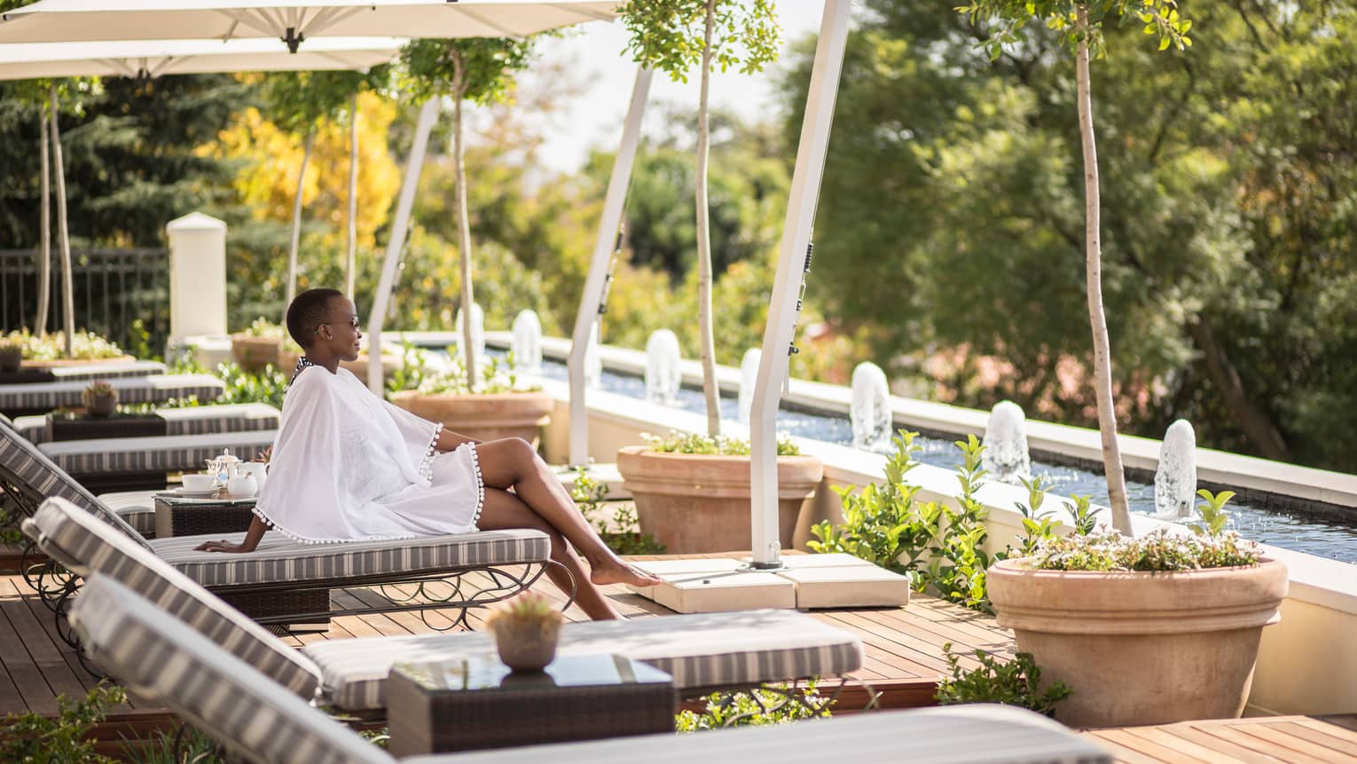 Woman in flowing white dress sits on lounge chair in row near outdoor swimming pool