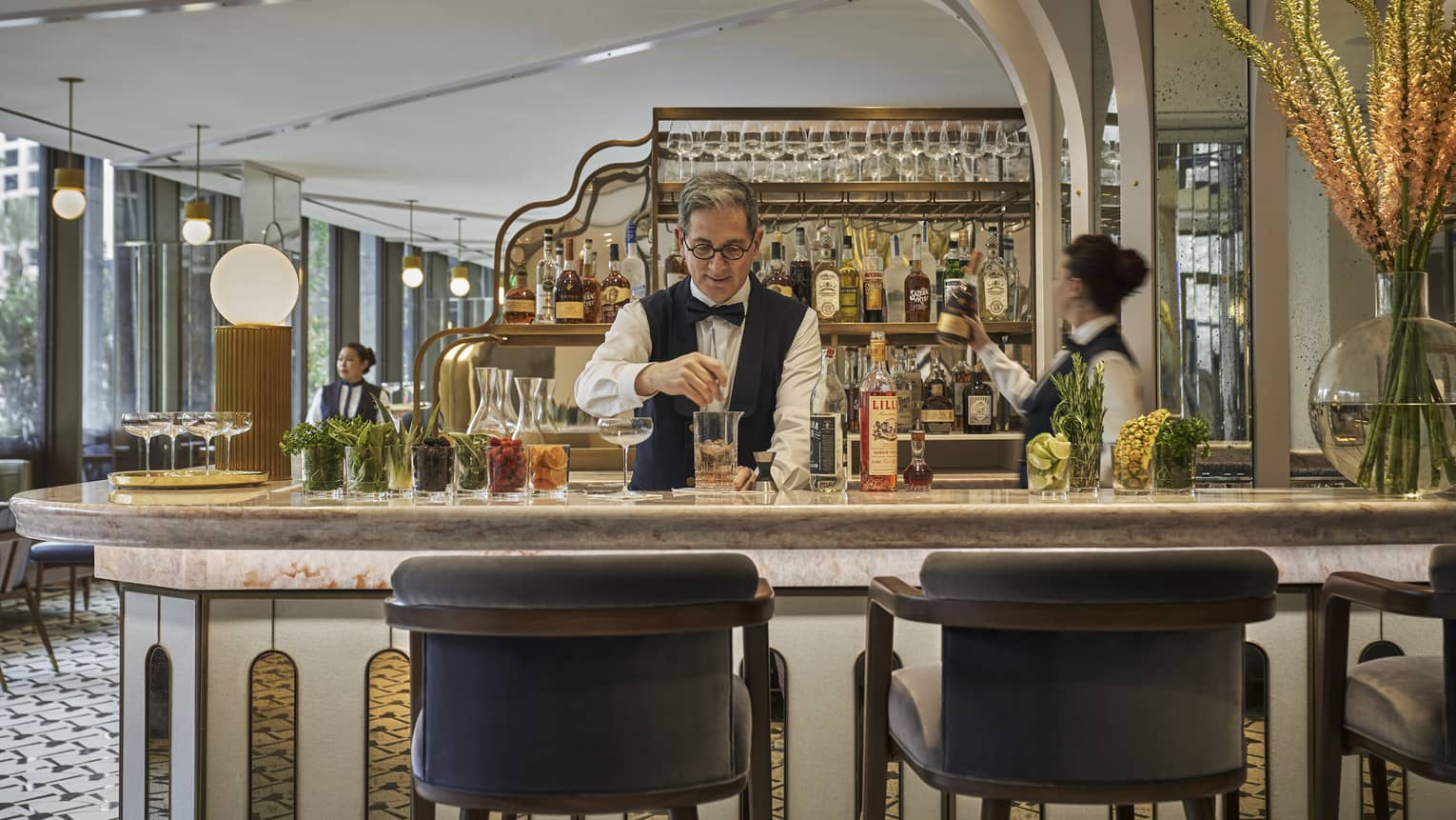 Two bartenders, one male and one female, prepare cocktails at Miss River's marble-topped bar, liquor bottles arranged behind them