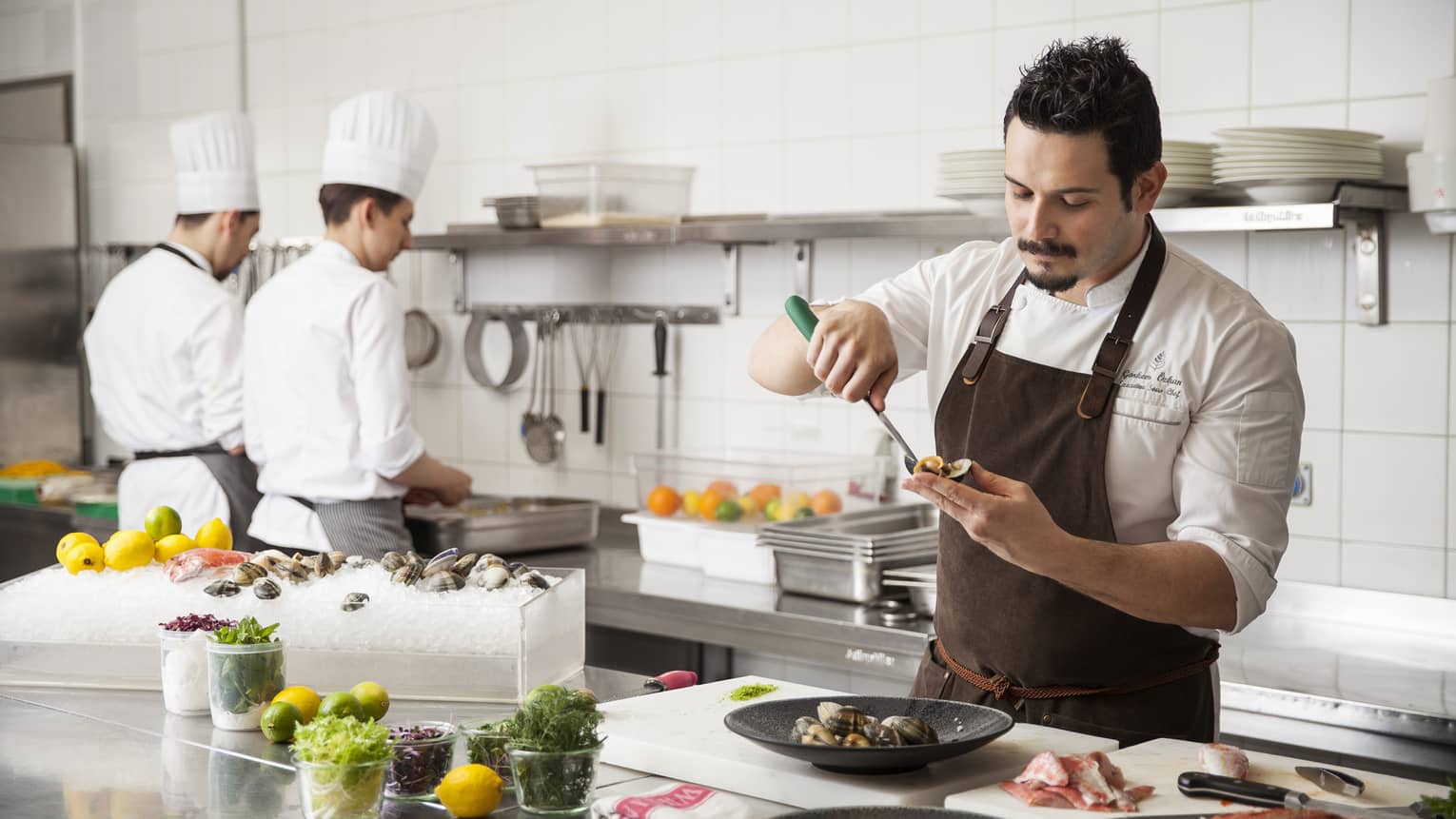 Chef Görkem Özkan garnishing oysters in restaurant kitchen, two cooks in background