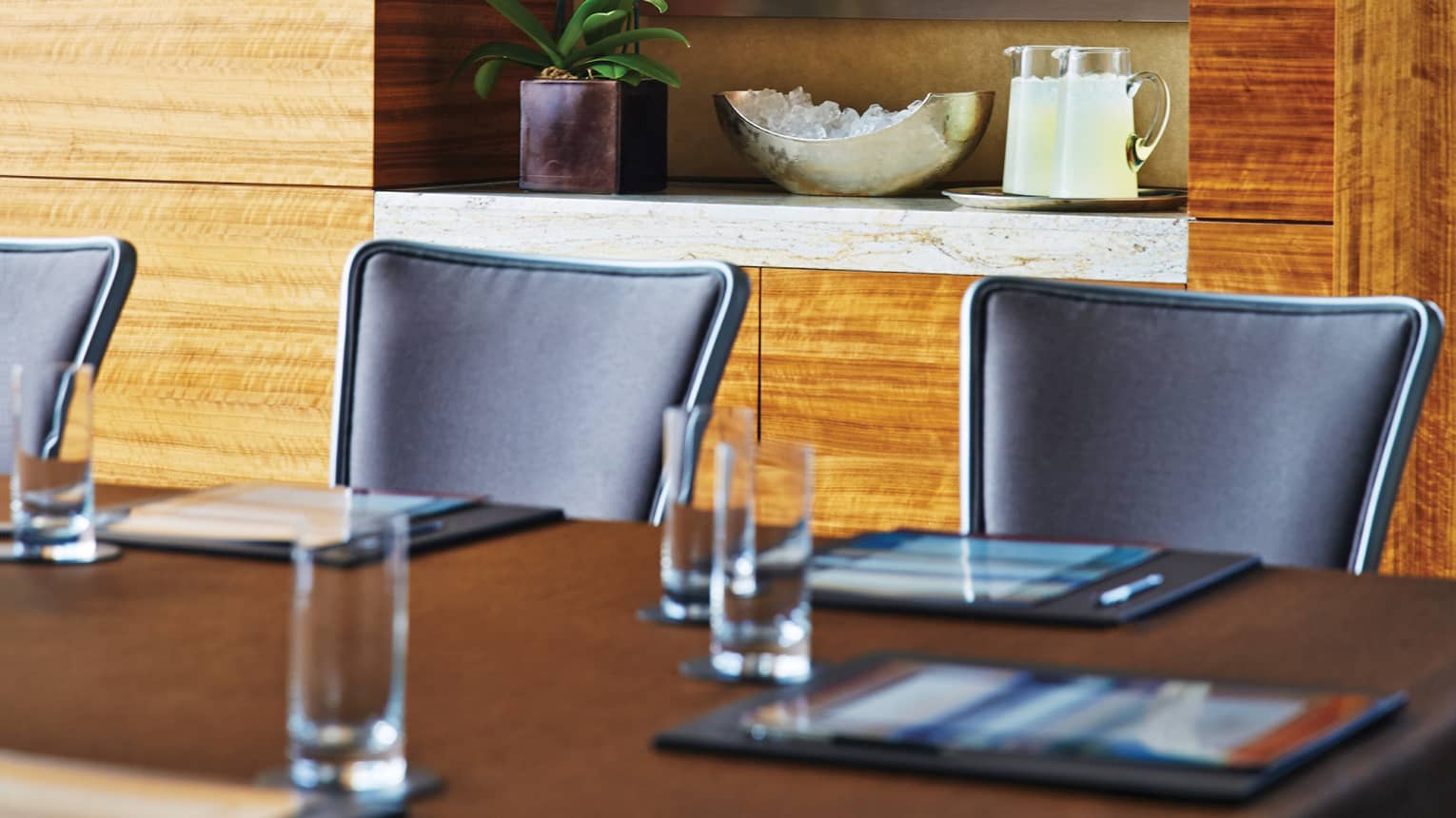 Boardroom table and black meeting chairs in front of modern wood wall, shelf with plant and jugs
