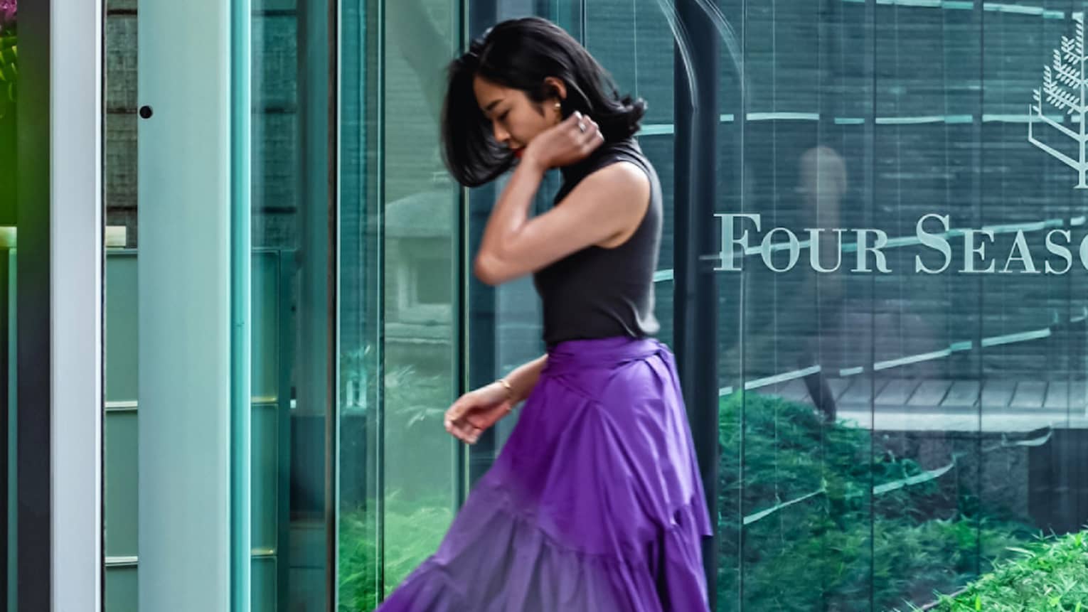 Person in a sleeveless top and purple skirt walking towards building with Four Seasons logo on glass door