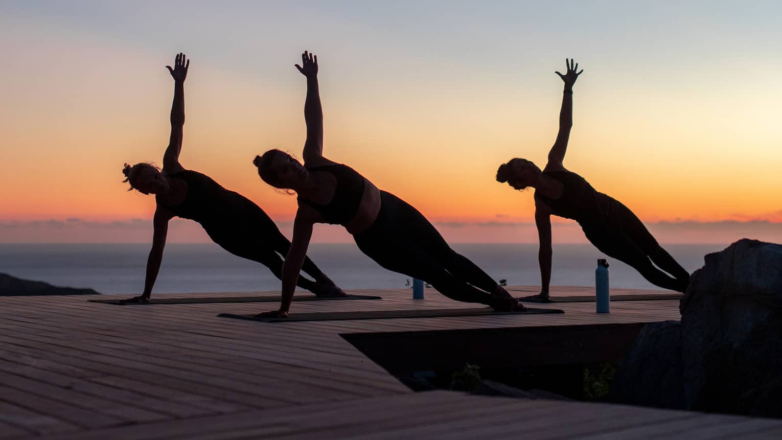 Three guests doing side plank poses on yoga mats on a wooden dock are silhouetted against a clear blue, orange and pink sky.