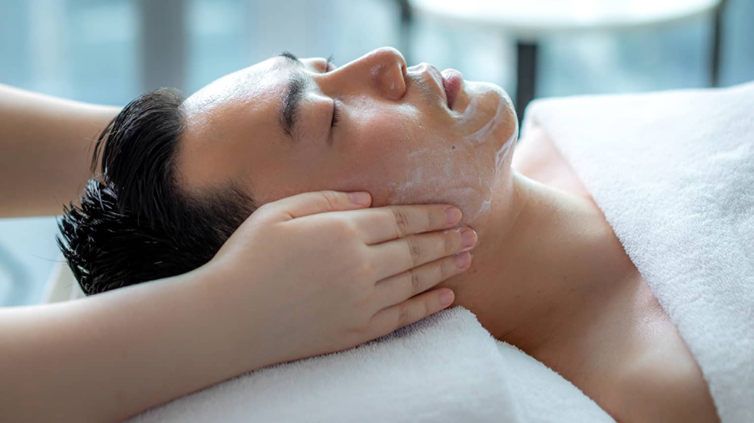 Hands rest on man's cheeks as he lays on massage table by glass window with daytime city view 