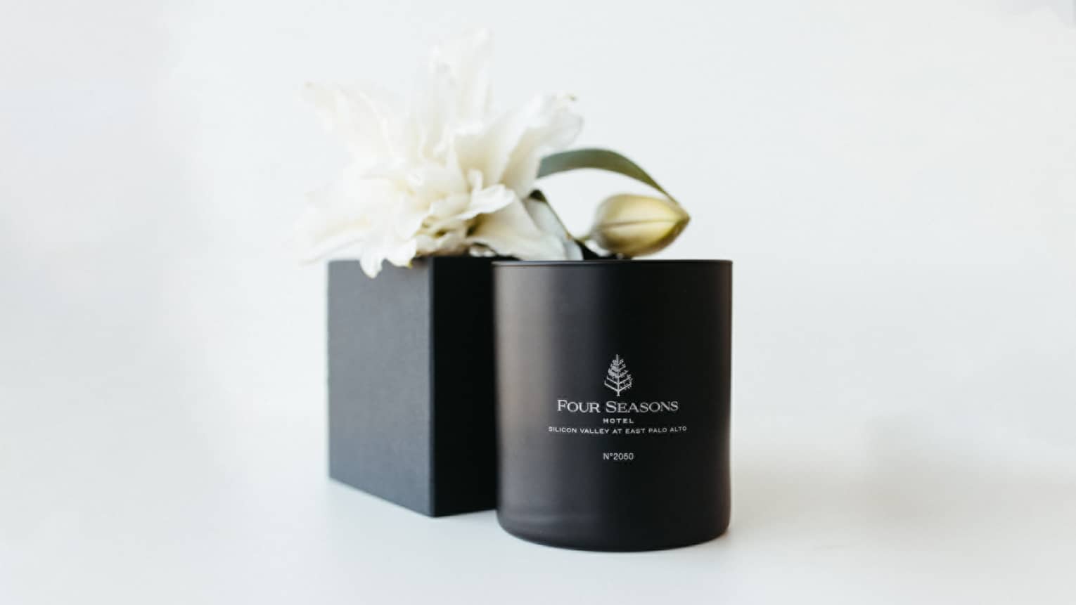 Black Four Seasons?branded candle placed next to a black box with a white flower on top, all on a white backdrop