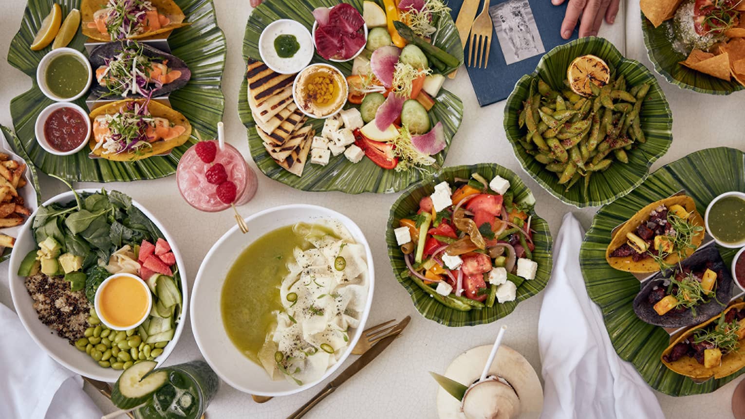 Assortment of vibrant dishes including salads, tacos, edamame, soup and tropical drinks served on a table with banana leaf platters