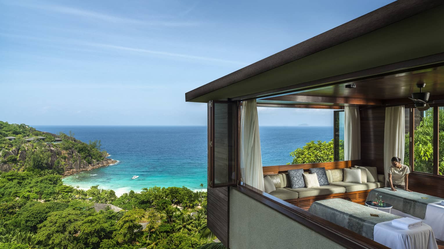 Woman smoothes sheet on side-by-side massage tables in spa room high above trees, ocean