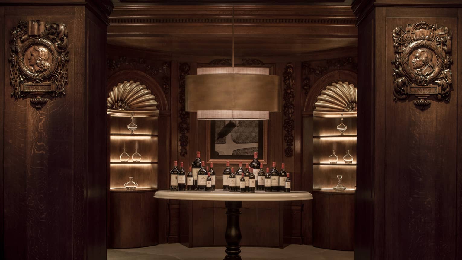 In a dark-panelled room with a large, stylish pendant lamp, a pedestal table holds twenty bottles of wine in varying sizes.