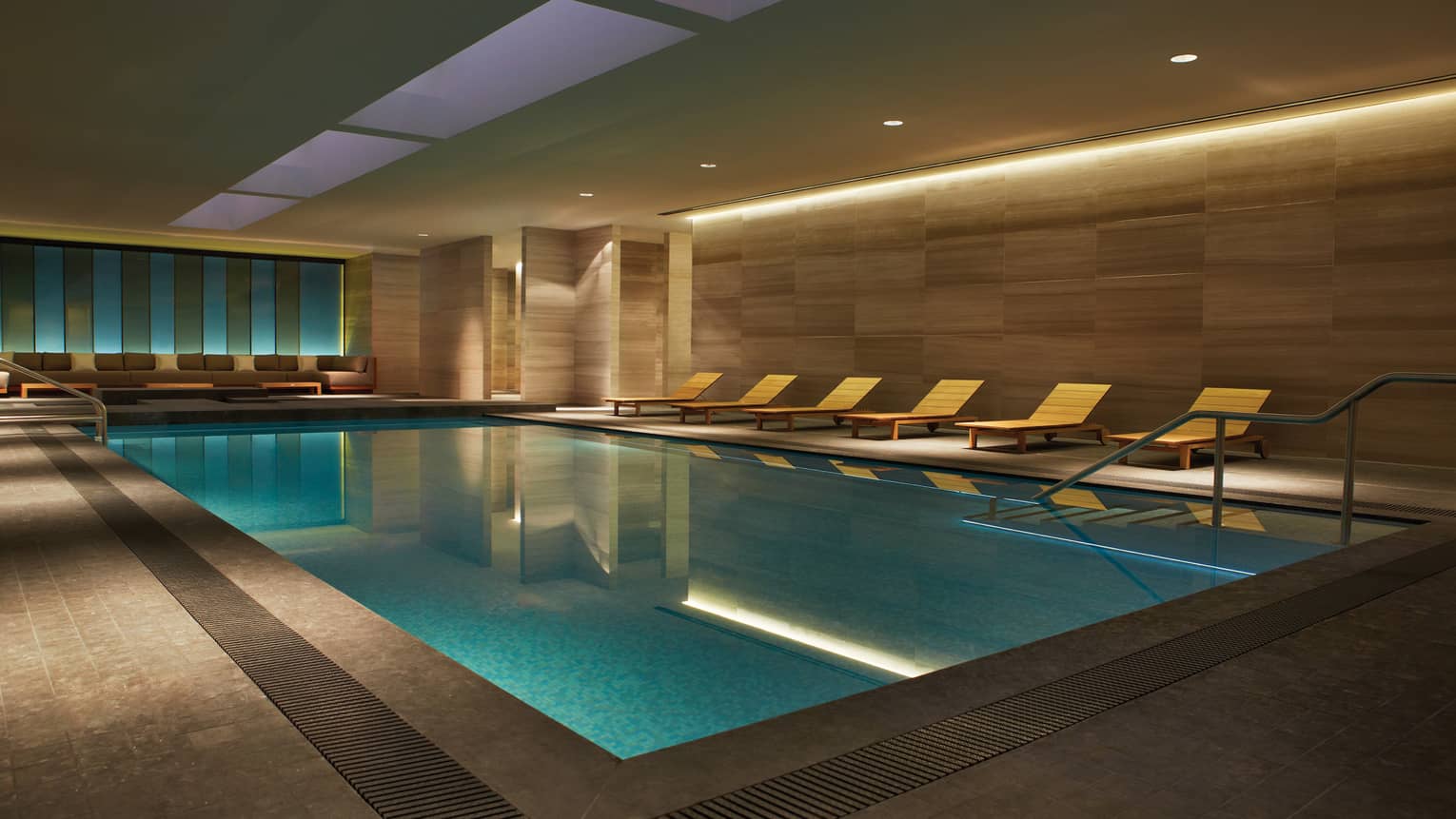 Illuminated indoor swimming pool with wood chairs on deck under granite walls