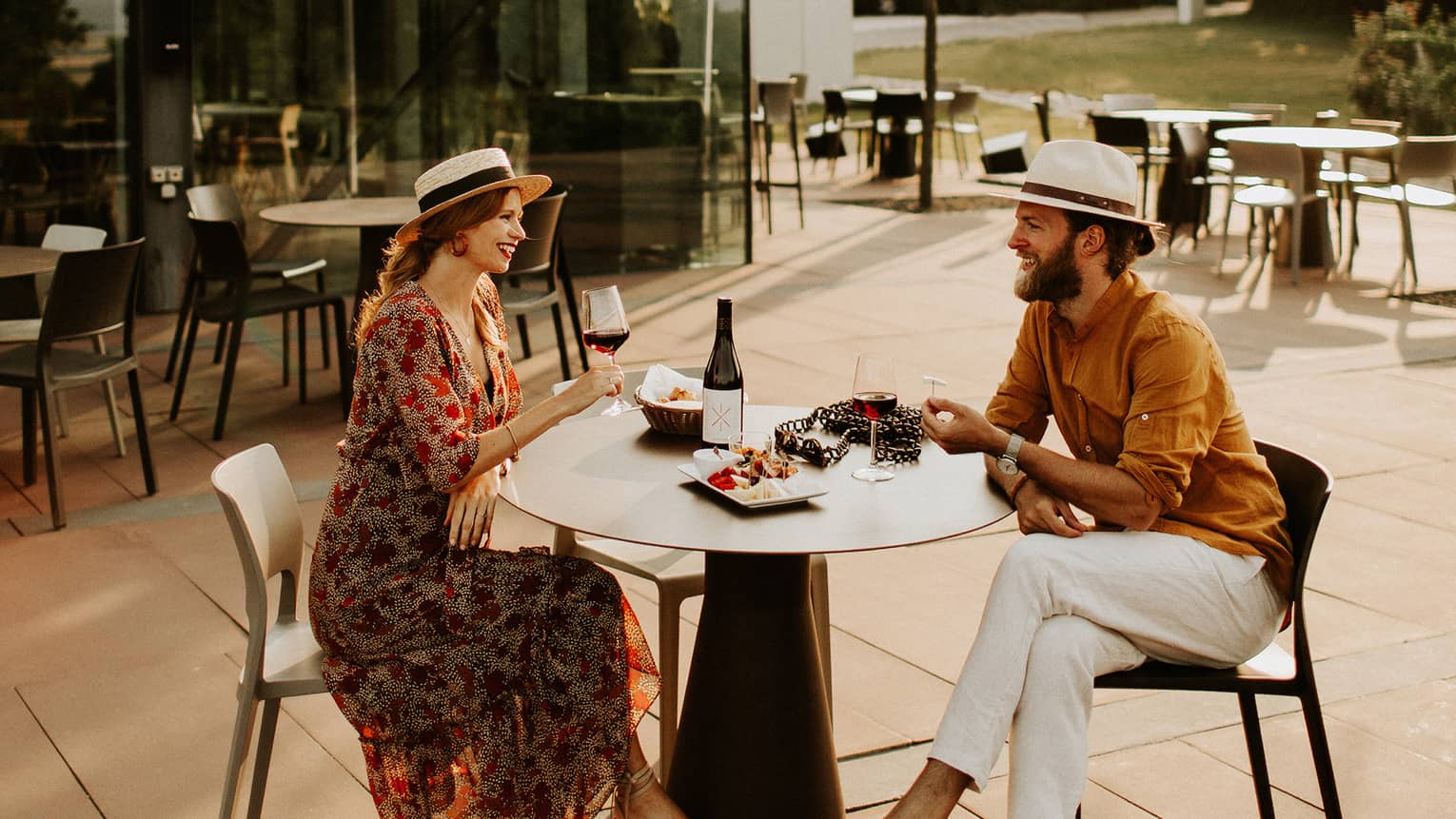 Couple wearing hats sit at outdoor table with plate of food, drinking red wine 