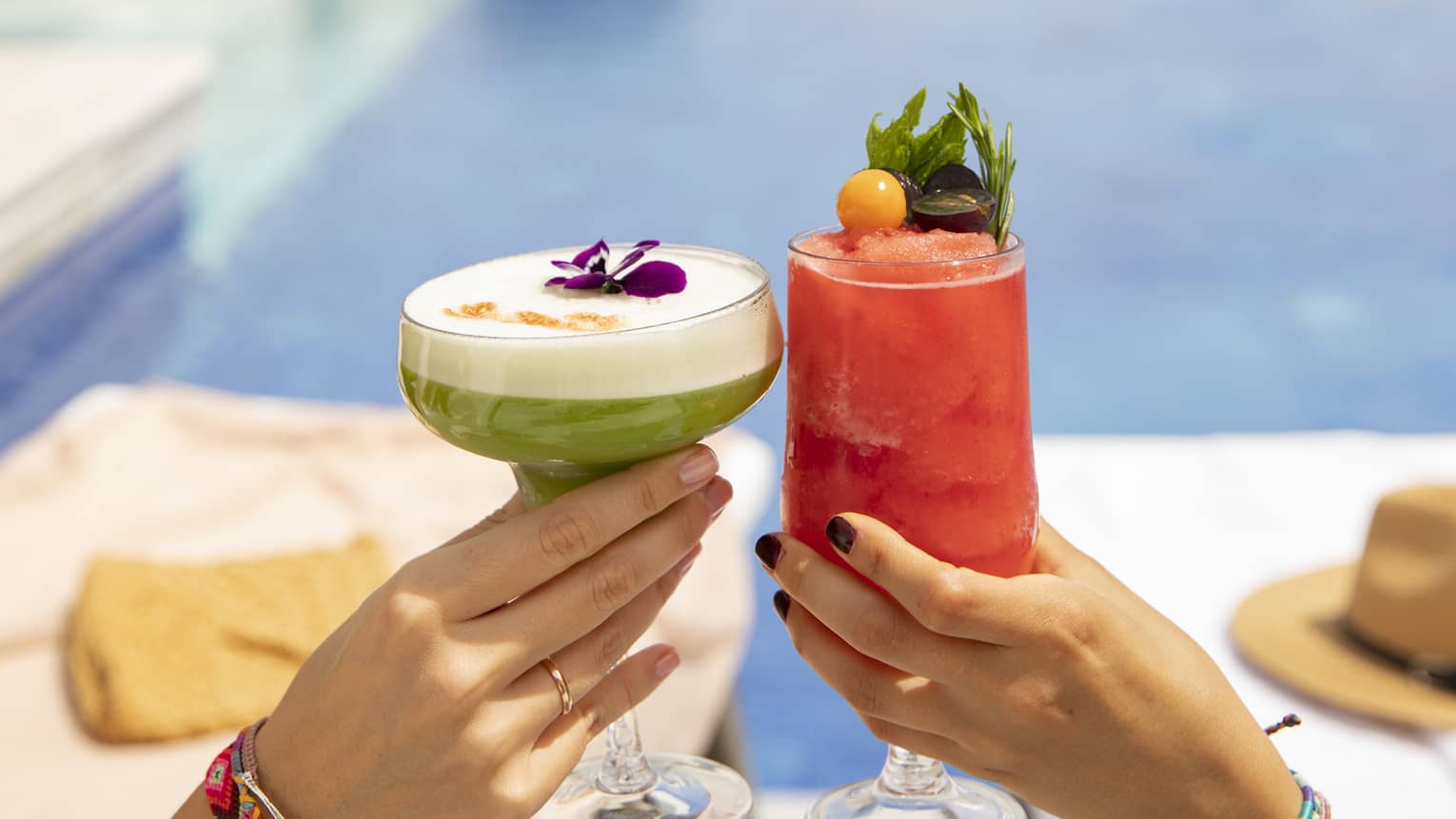 One hand holding white-topped green cocktail toasting a hand holding pink cocktail poolside