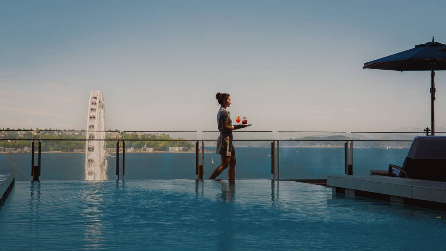 A woman carries a tray of beverages next to infinity pool overlooking city harbour