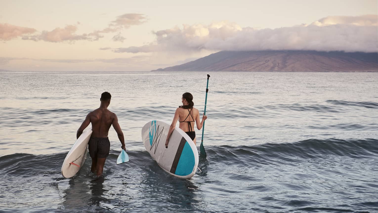 Two people standing in the ocean holding paddleboards with mountains in the background