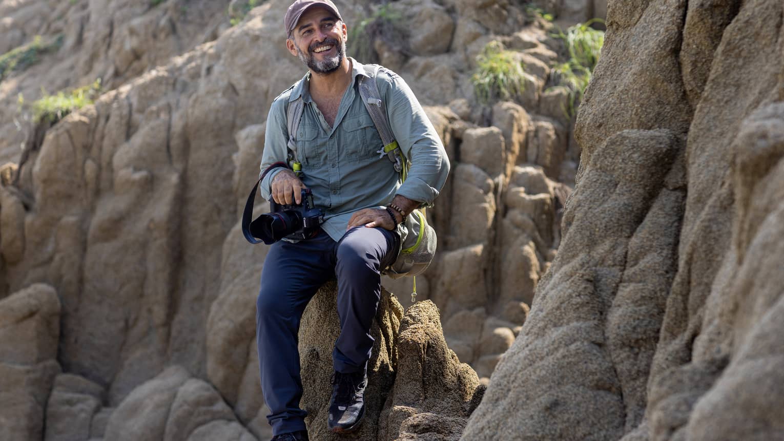 A photographer wearing a backpack and holding a camera in one hand sits atop a rock formation, smiling into the distance.