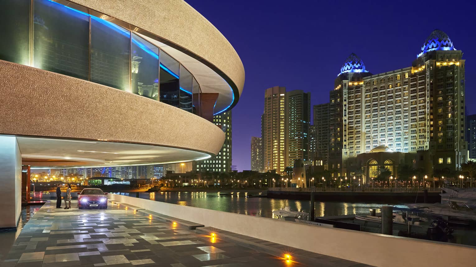 Luxury car with headlights on parked under curved Nobu restaurant exterior at night, city lights