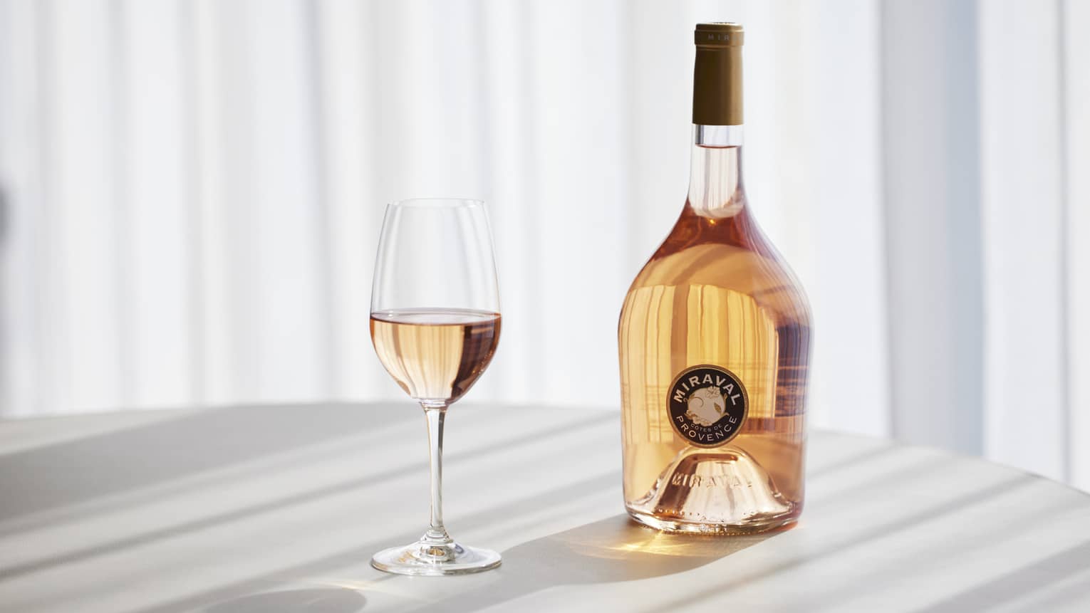 Miraval cocktail in stemmed glass next to corked bottle of liquor, sunlight streaming into to create shadows