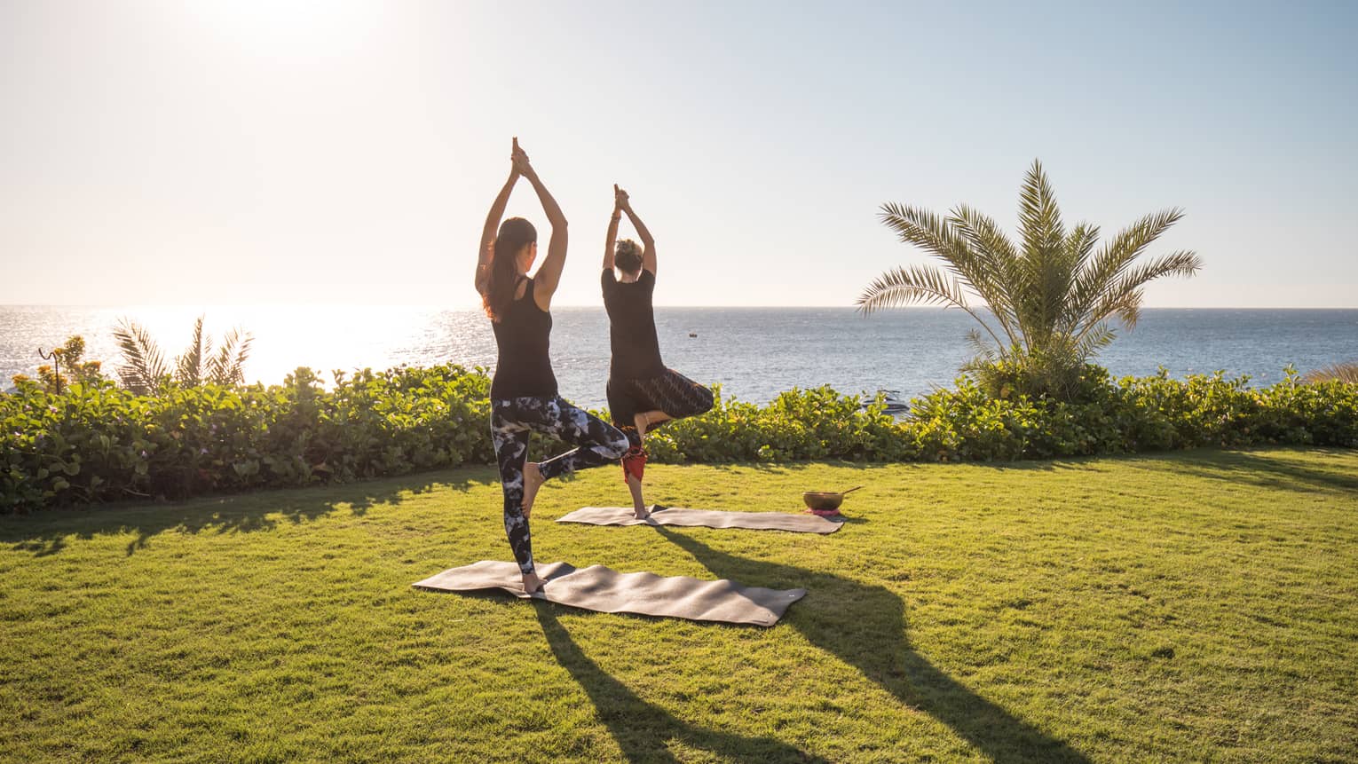 Two people enjoy yoga outdoors by the sea with the sun rising in the sky at a luxury hotel