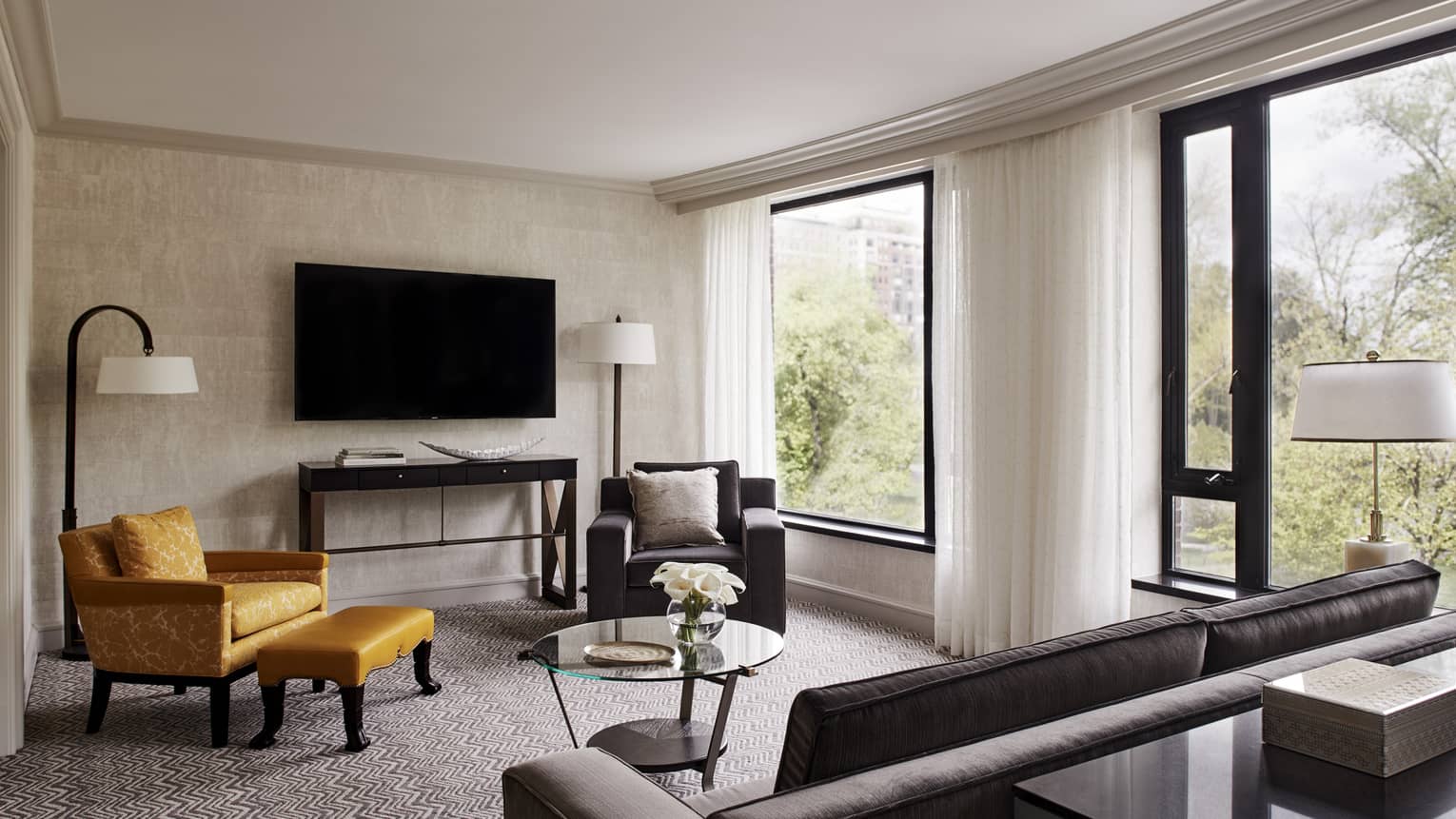 A living room with a brown sofa, yellow arm chair and ottoman, brown arm chair, TV, windows