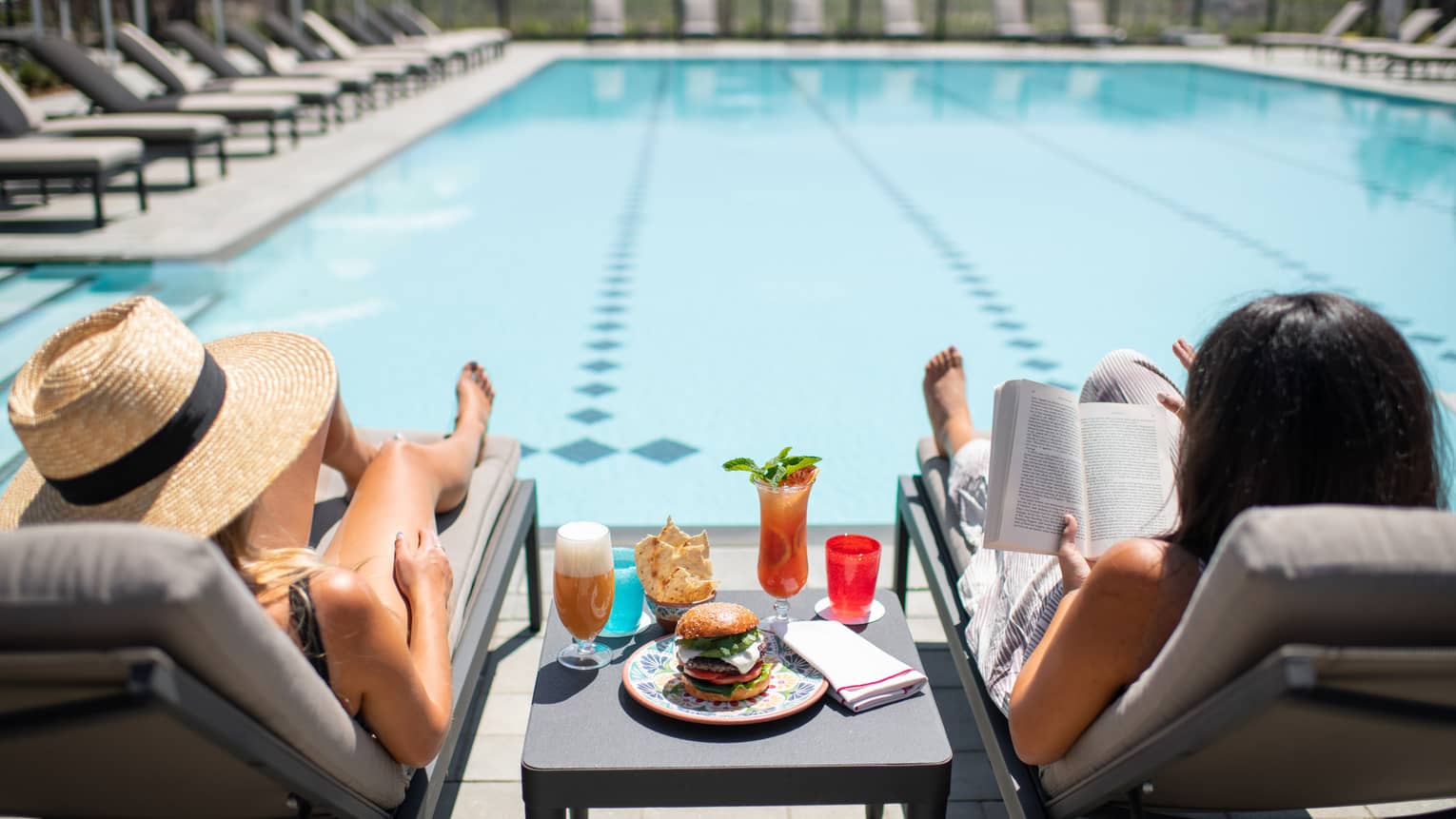 Two women sitting on lounge chairs next to a pool with food and drinks.