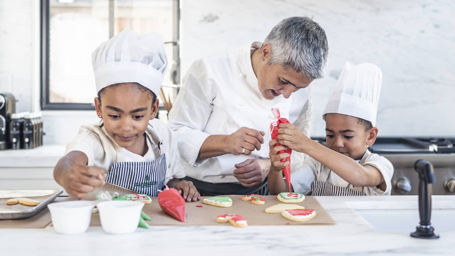 Two children in chef’s hats and aprons in a stark, white kitchen; a chef watches closely as one pipes icing onto a cookie.