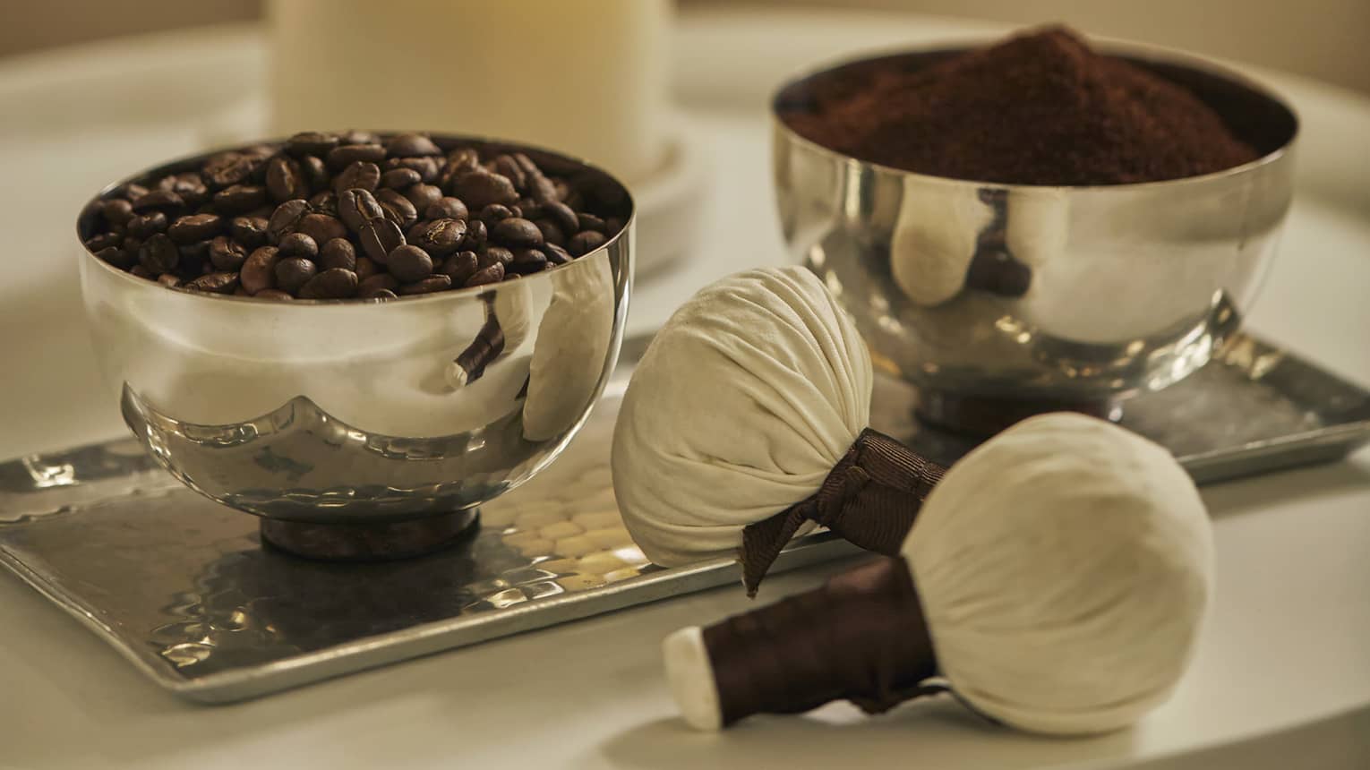 Two silver bowls on tray hold roasted coffee beans, ground coffee powder