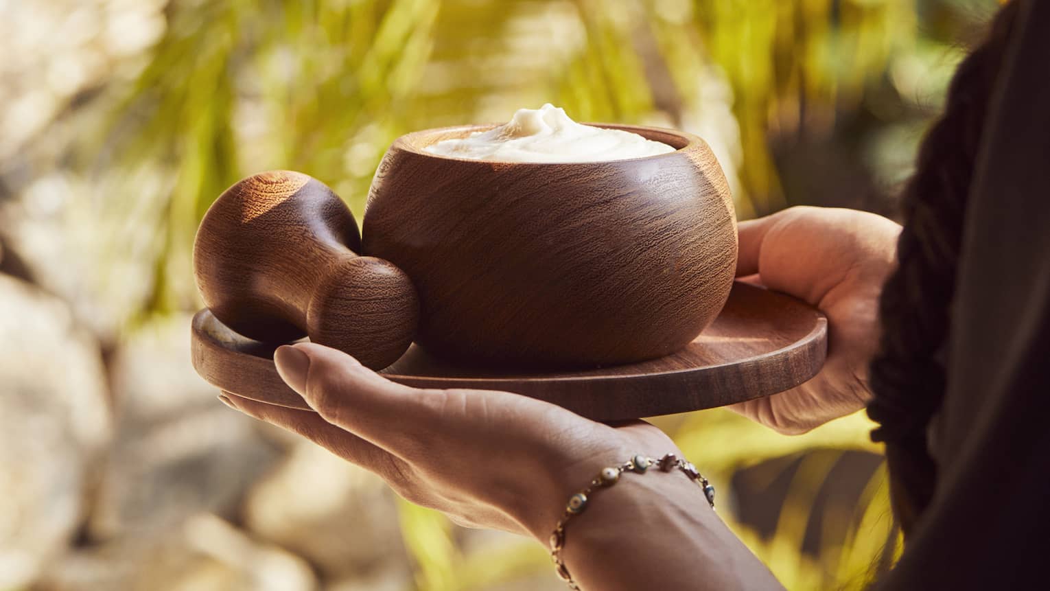 Two hands carry a small round tray with a wooden pestle and a bowl filled with a creamy body scrub past golden palm fronds.