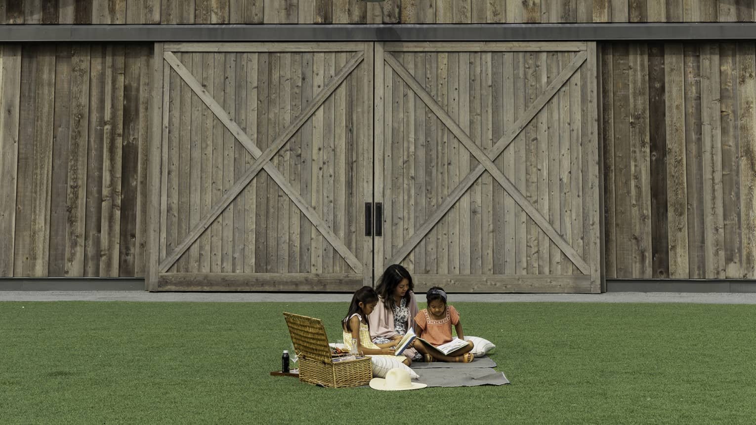 A family sits and reads on a picnic blanket with a basket on a vast expanse of green grass in front of a large wooden barn.