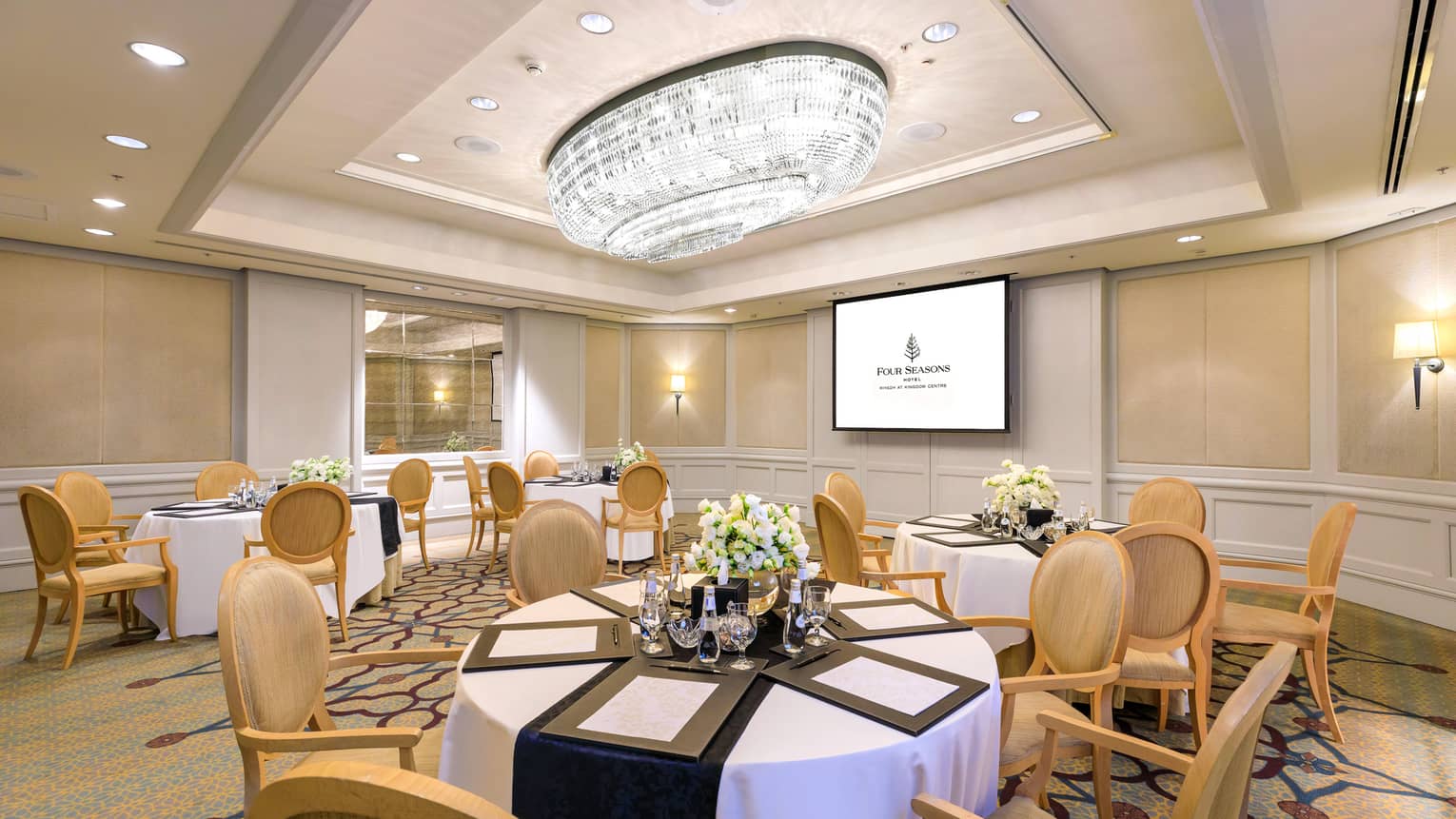 Elegant hotel meeting room with six banquet tables, presentation screen and crystal chandelier