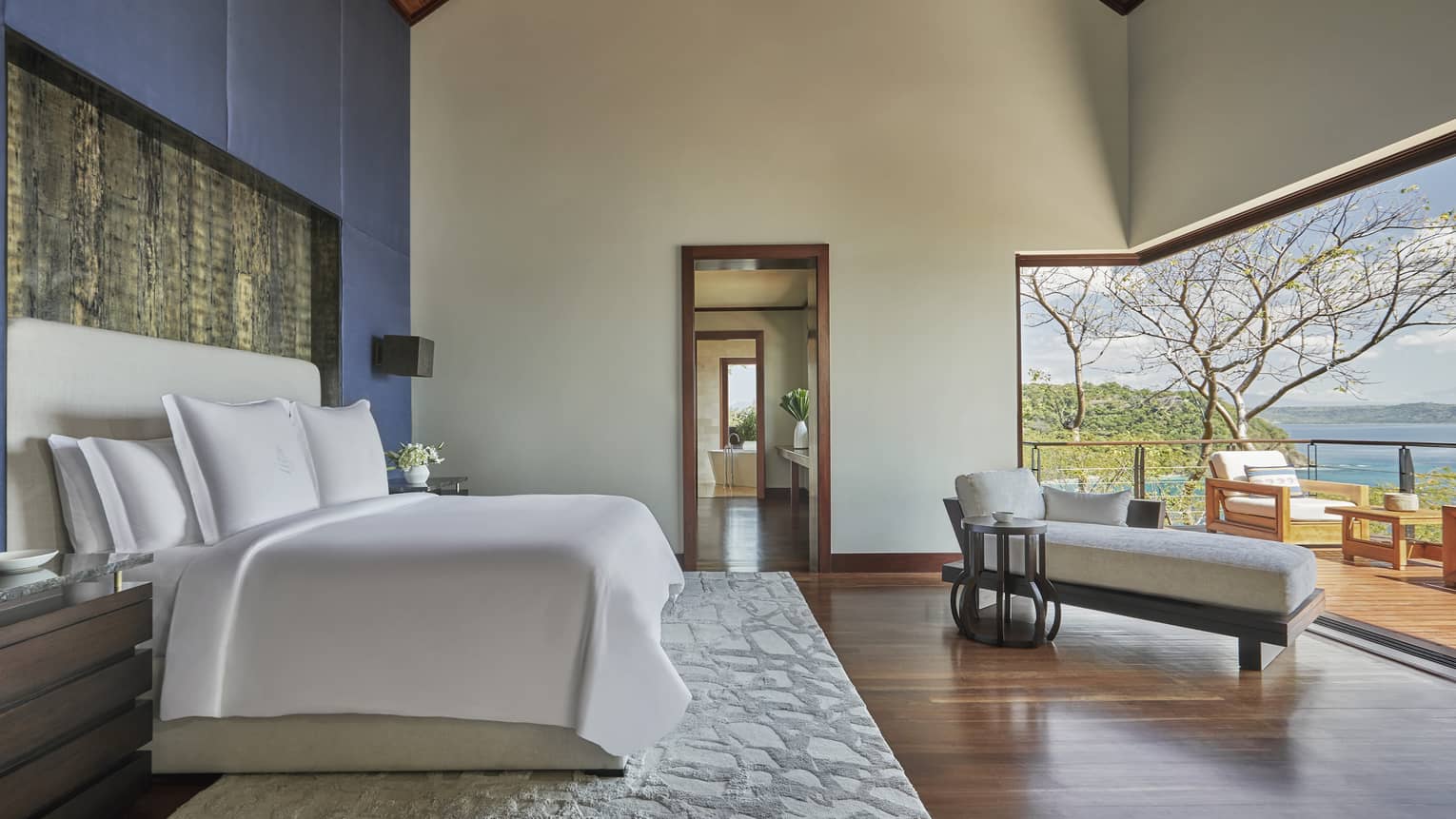 Bedroom with blue accent wall, wooden floors, rug, chaise, opening to terrace