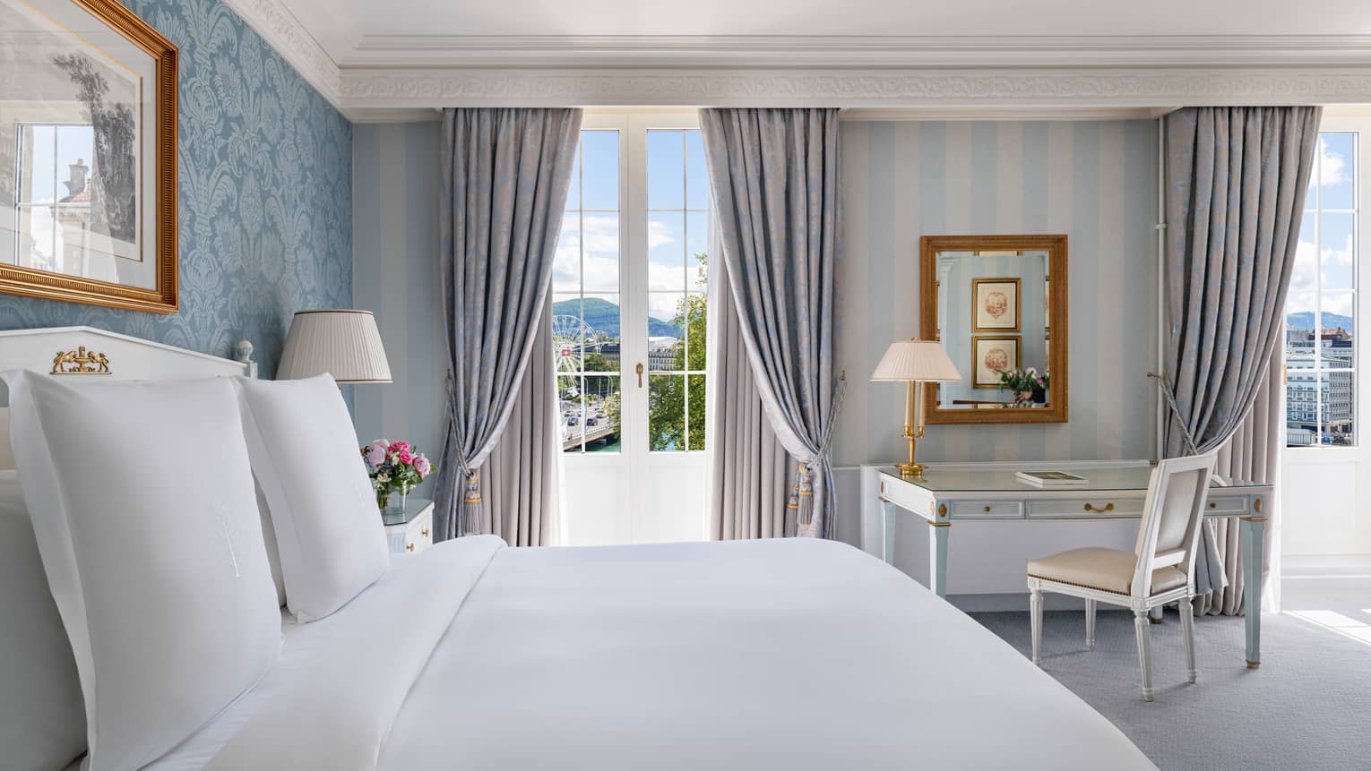 Luxury hotel room with a king bed, floral and striped wallpaper, glass-top desk, floor-length curtains and a window overlooking a ferris wheel and water
