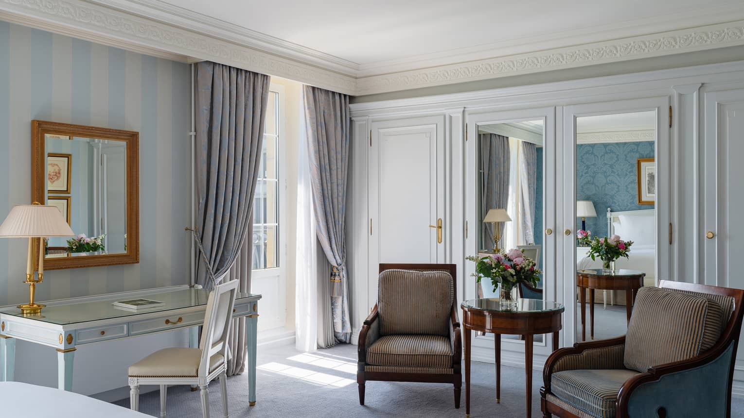 Luxury hotel room with a glass-top desk, upholstered armchairs, mirrored wardrobes and floral arrangement