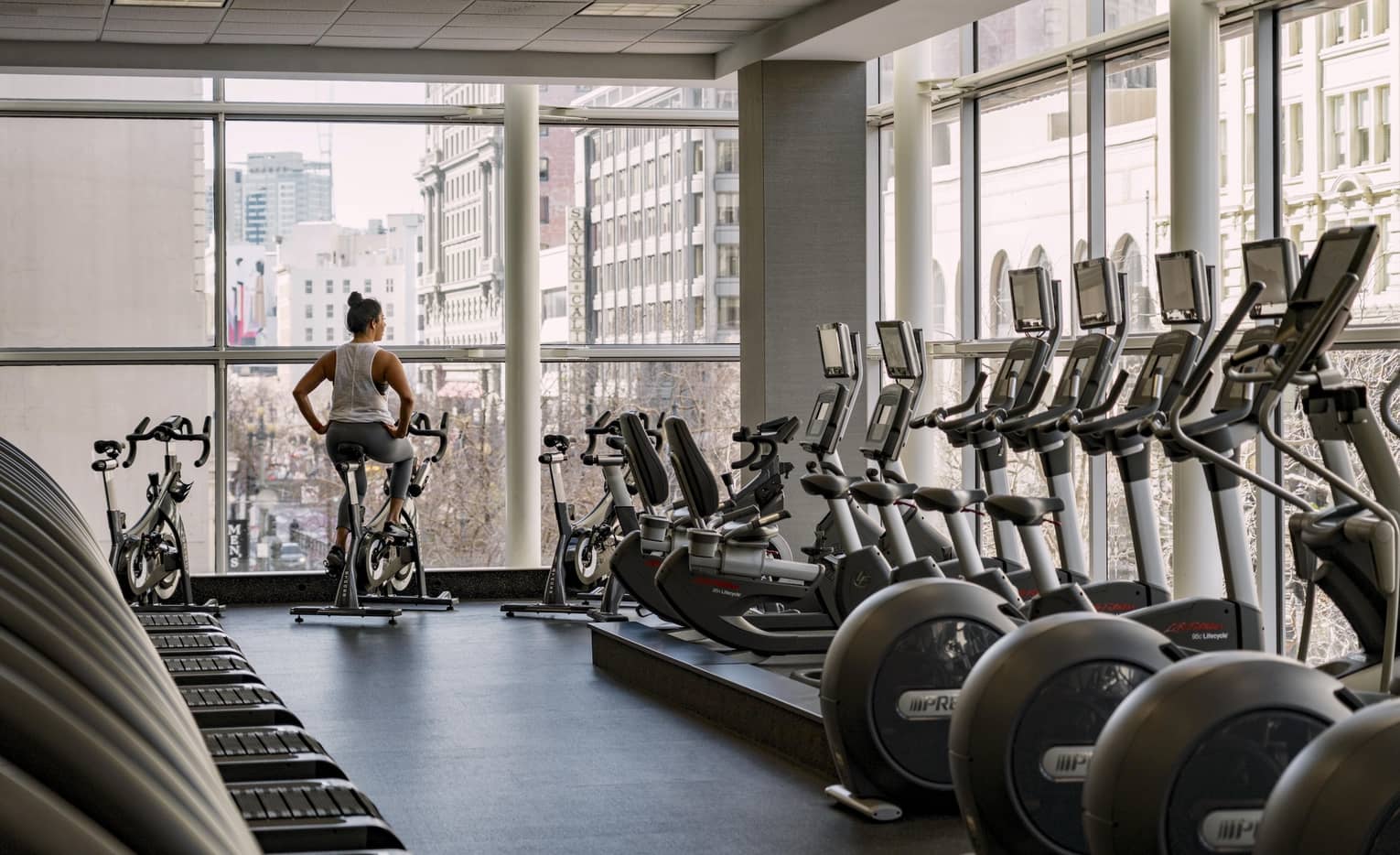 A woman rides a stationary bike in a hotel fitness center with floor-to-ceiling windows overlooking a city view