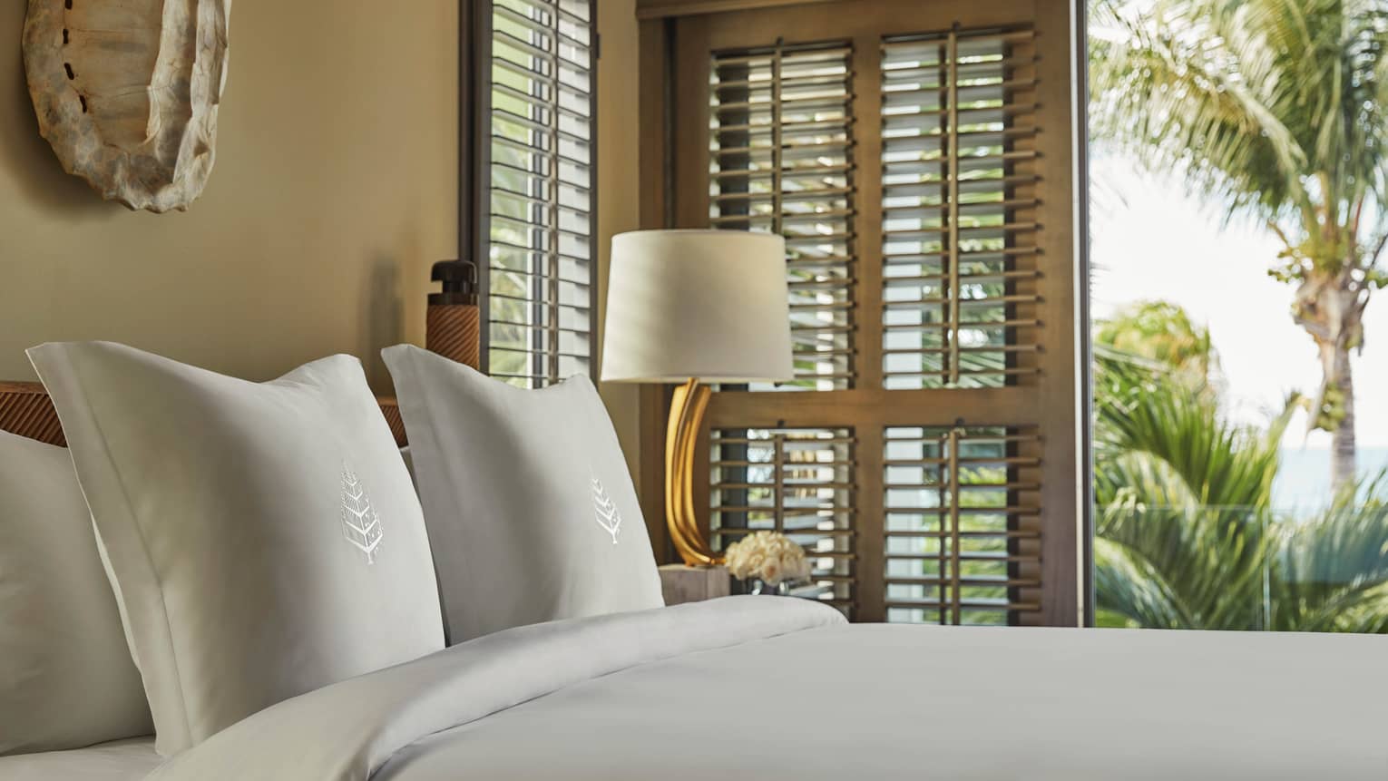 Close-up of a neatly made bed with embroidered white pillows, next to a lamp and window shutters revealing tropical palm trees outside