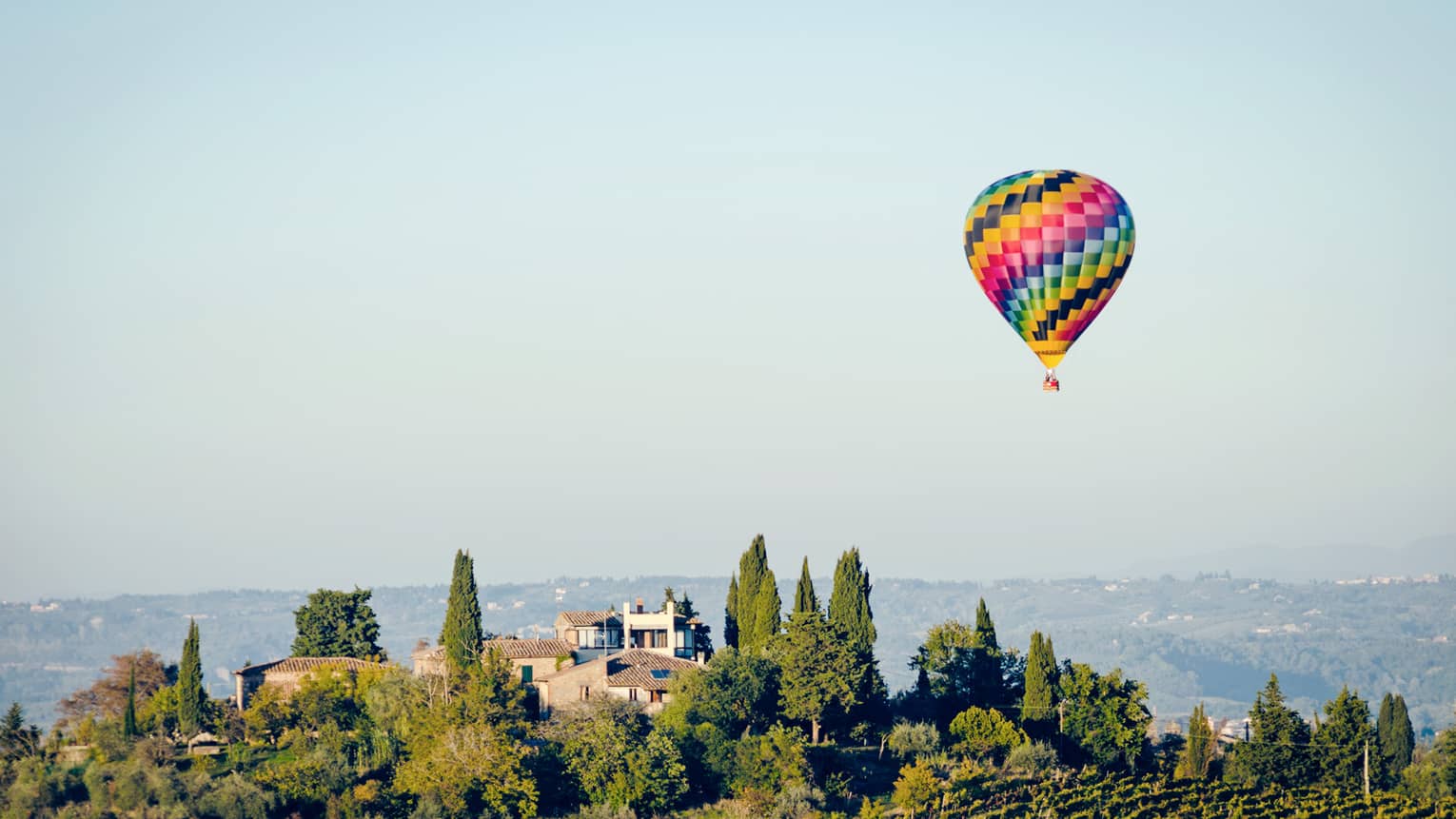 Colourful hot air balloon floating above a landscape with trees and houses, set against a clear blue sky.		