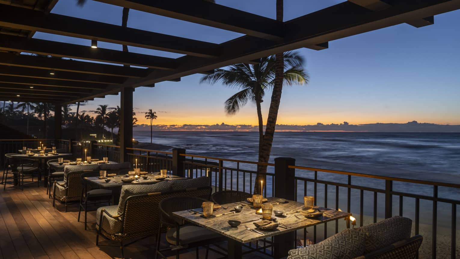 An intimately lit outdoor dining space with the ocean, palm trees and a dramatic sunset in the background
