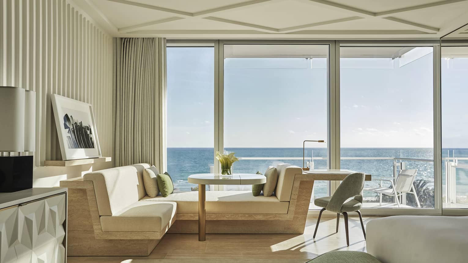 A hotel room with large open windows looking out at the ocean with a chaise lounge, and a small table and chair.