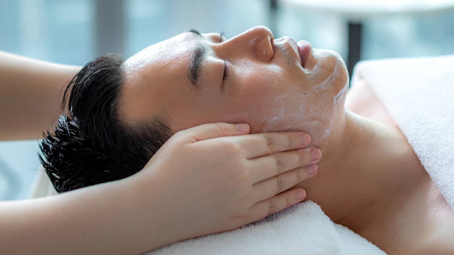 Hands rest on man's cheeks as he lays on massage table by glass window with daytime city view 