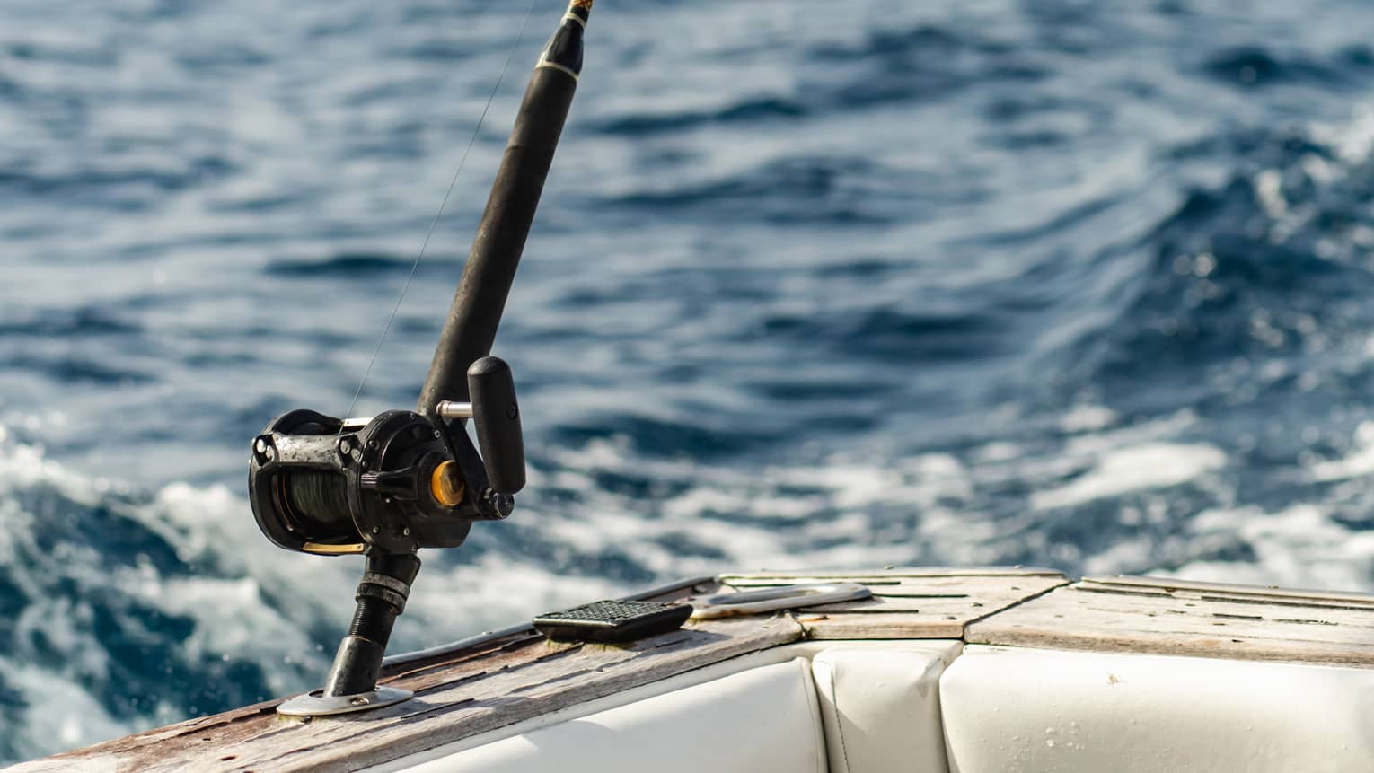 Close-up of a fishing rod secured in a holder at the back corner of a boat on an expanse of wavy water under pale blue sky.