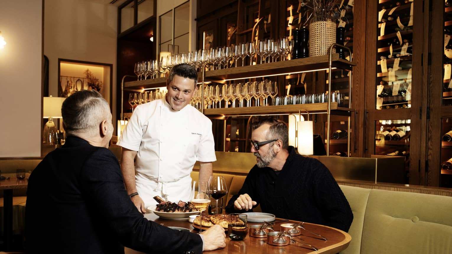Two guests sitting at a dining table in a restaurant while the chef stands near the table talking to them.