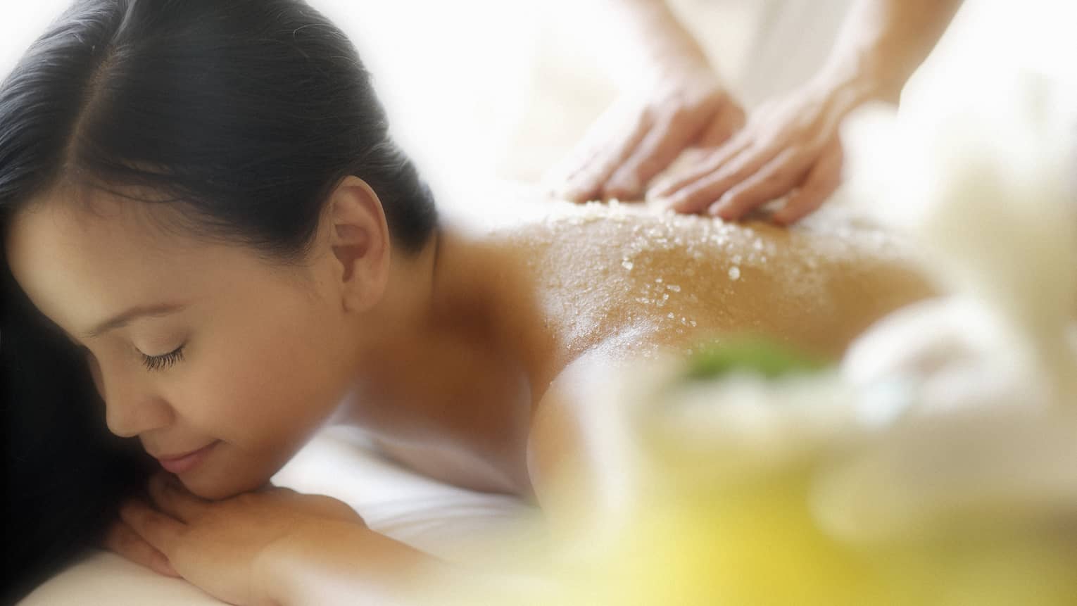 Woman lying down with eyes closed as hands massage salt scrub into shoulders