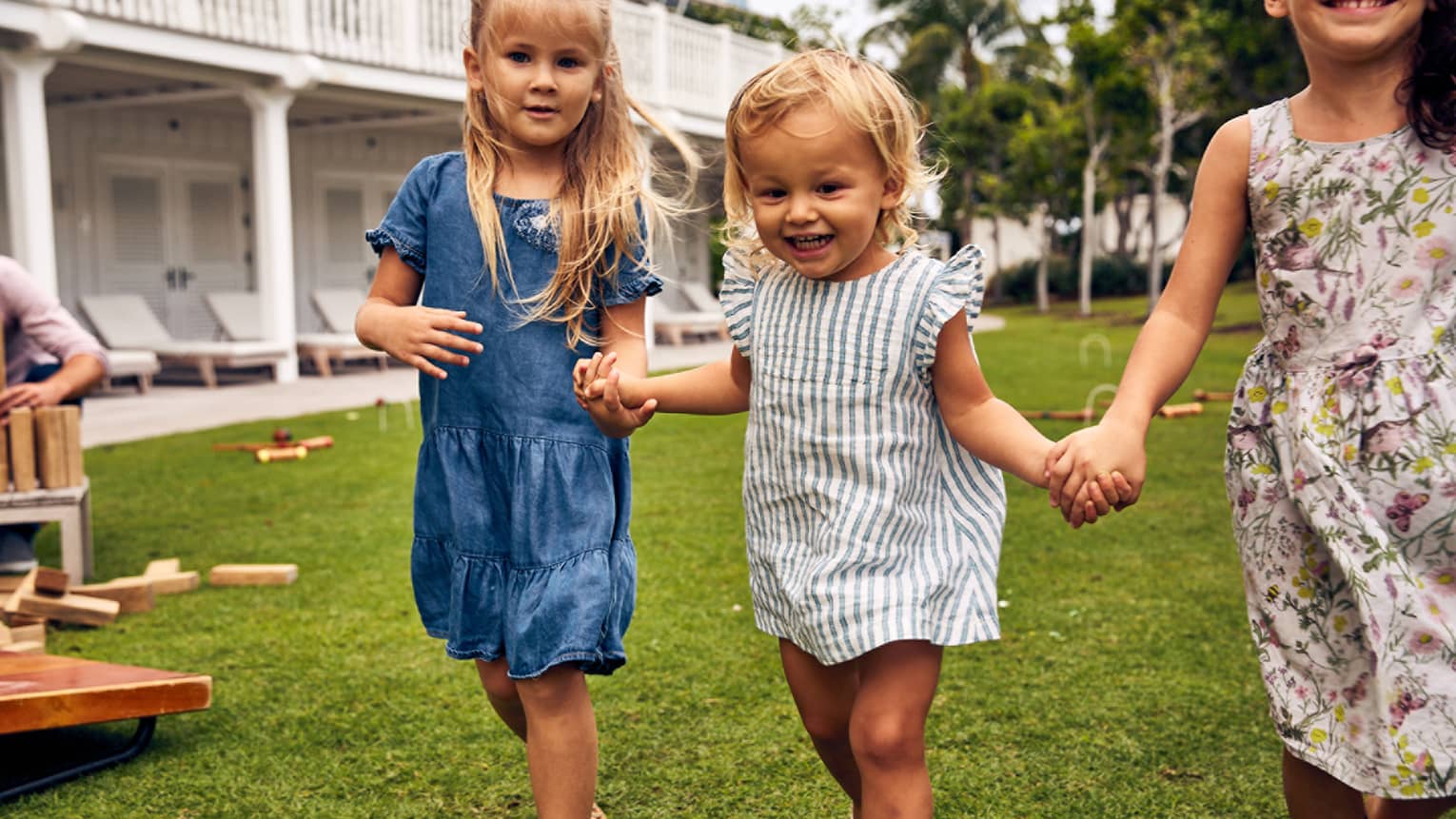 Three young girls holding hands as they play on grass next to a large white building.